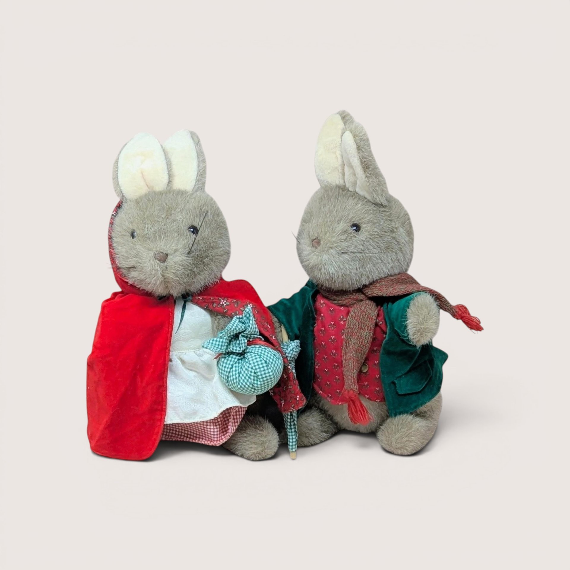 Two plush rabbit dolls dressed in vintage cloaks and winter clothing, centered on a beige background.