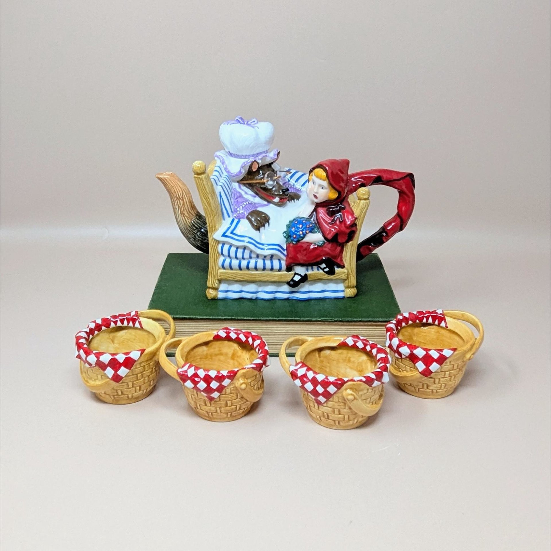A colorful Dept 56 ceramic teapot set showing Little Red Riding Hood sitting by the Big Bad Wolf in a bed, accompanied by four small tea cups shaped like wicker picnic baskets