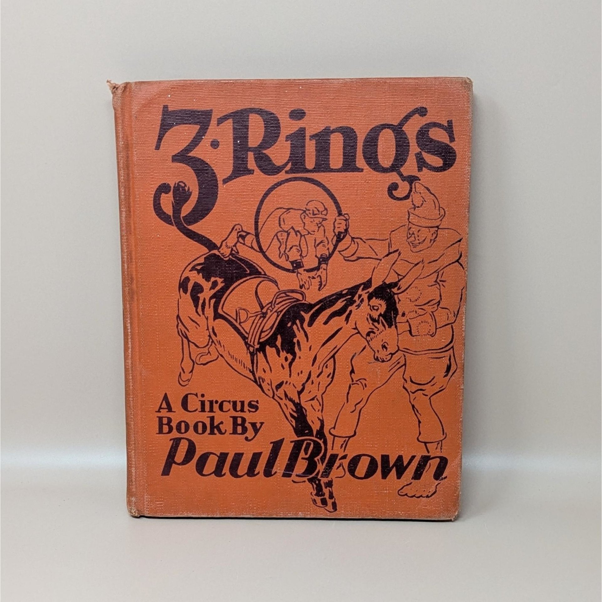 A vintage 1938 hardcover book titled 3 Rings A Circus Book by Paul Brown with an orange cloth cover featuring a black line drawing of a circus clown and donkey