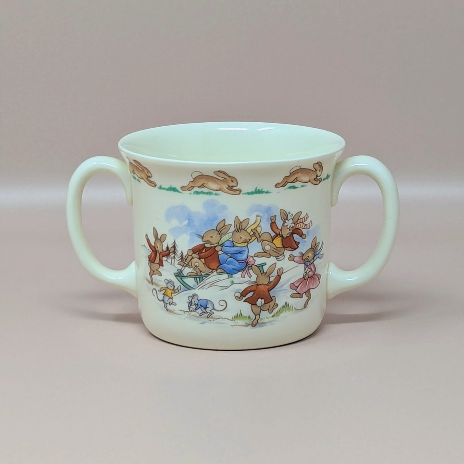 A vintage cream colored two handled mug featuring a group of rabbits on a sled with a border of running rabbits around the rim