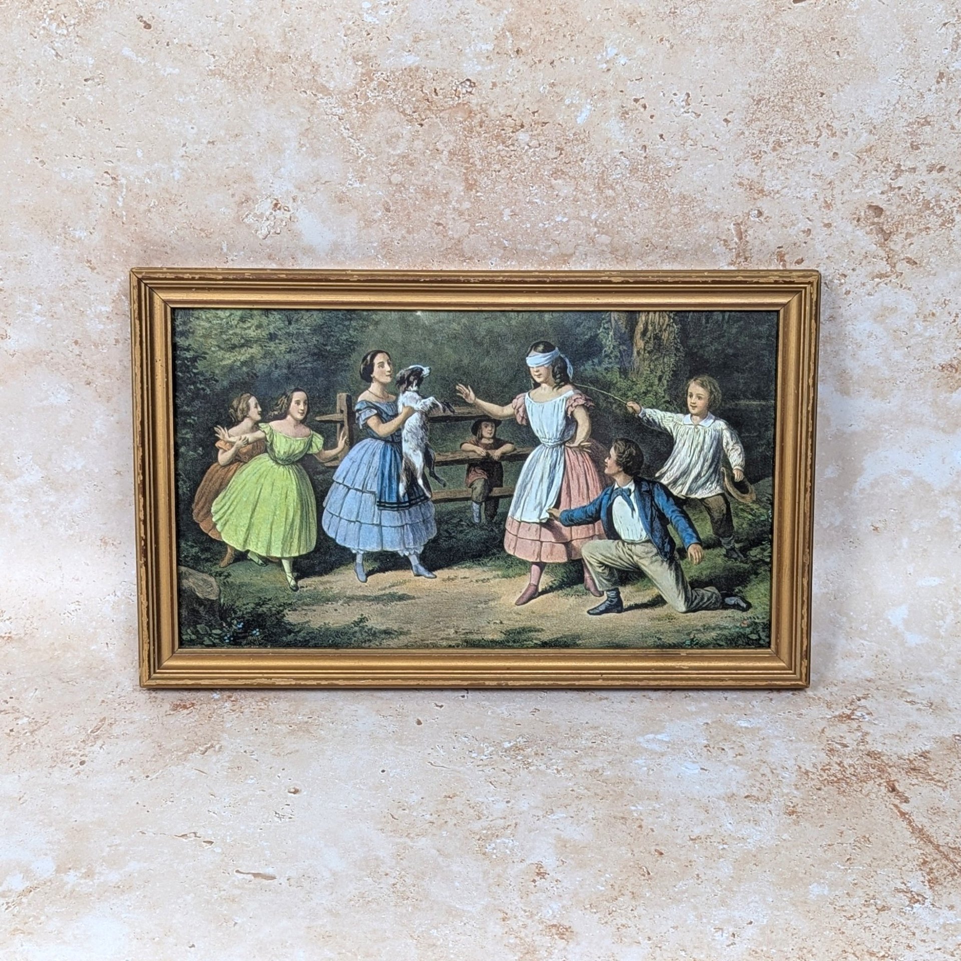A vintage framed Currier and Ives print titled Blind Man's Buff on a tan textured background. The mid-century horizontal lithograph depicts Victorian-era children playing a blindfolded game in a lush green backyard space.