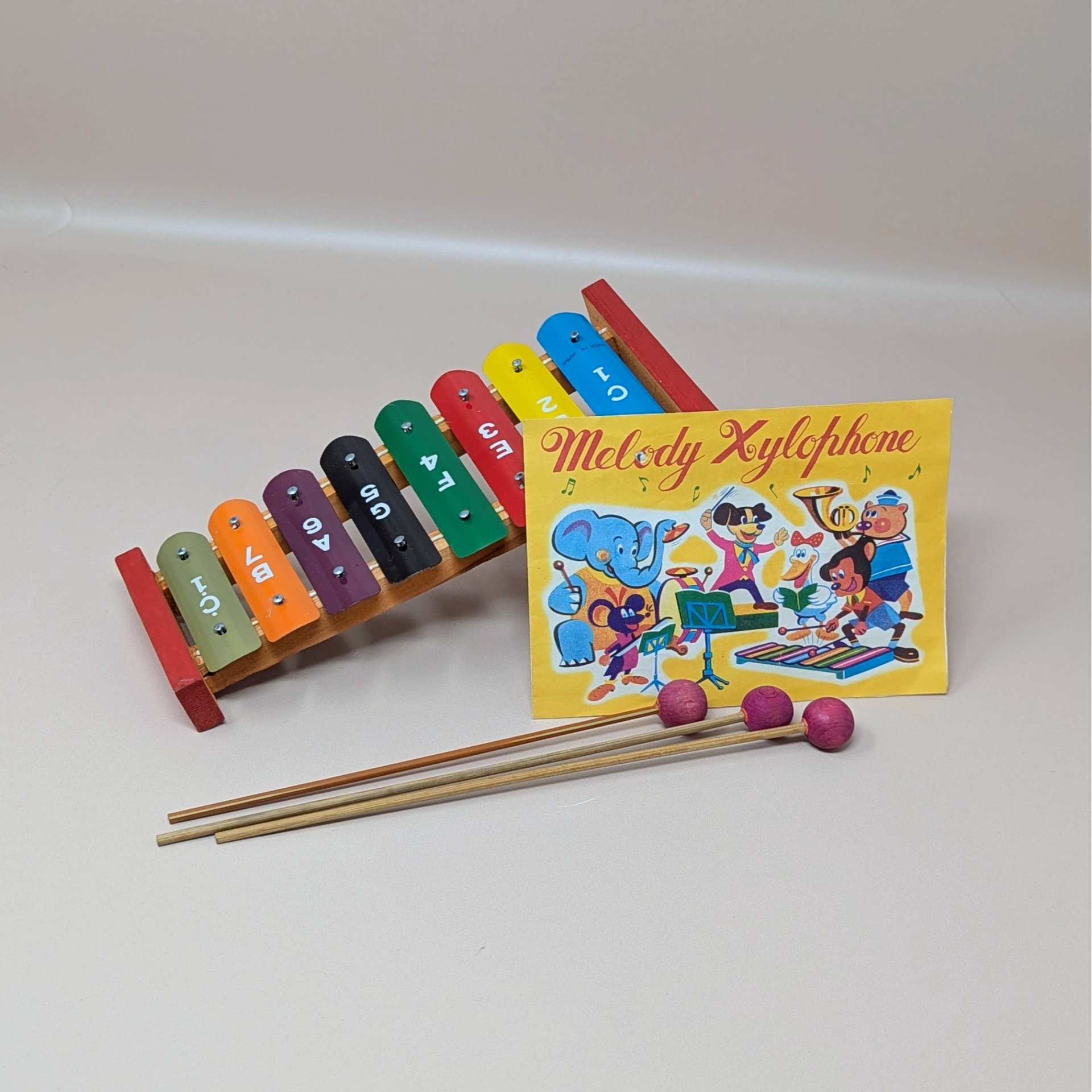 A vintage colorful metal xylophone with eight rainbow colored keys alongside three wooden mallets and a yellow music instruction sheet featuring cartoon animals