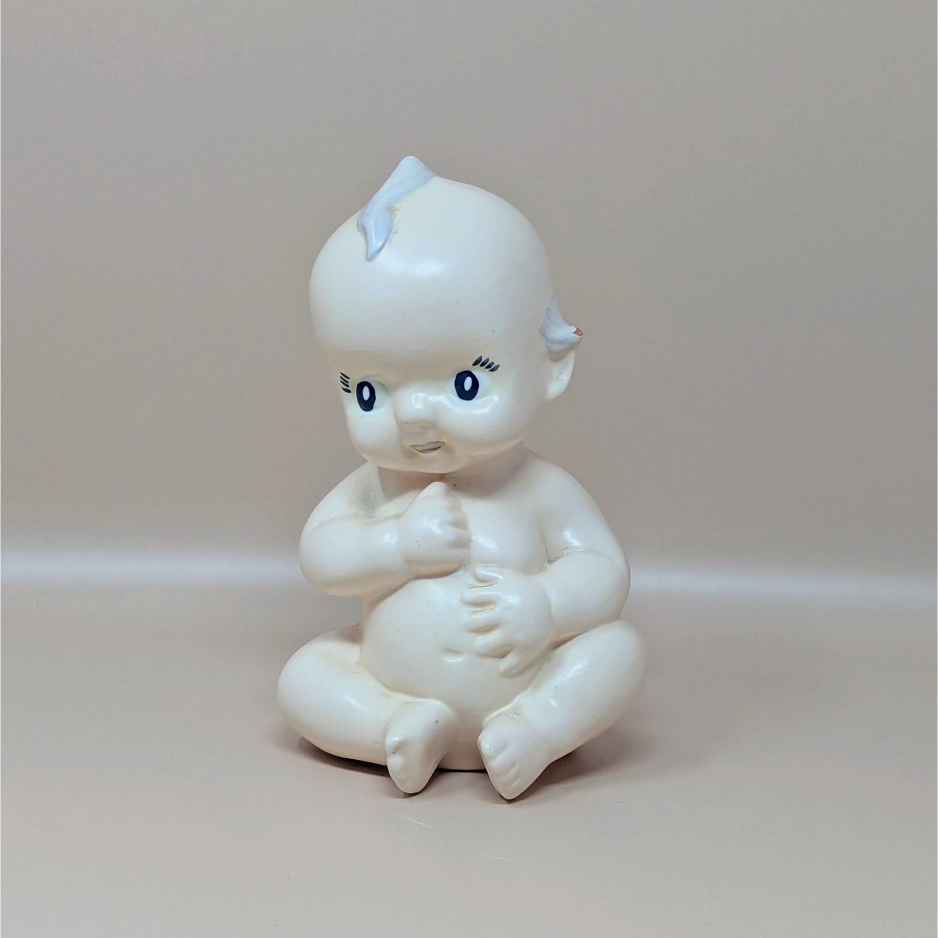 A vintage white ceramic Kewpie baby doll figurine sitting cross-legged with hand-painted blue side-glance eyes and small blue wings