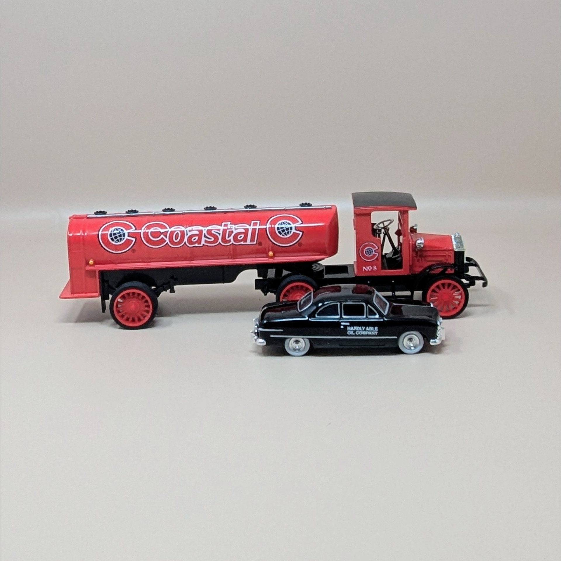 A red vintage-style 1920 Pierce Arrow tanker truck with Coastal Oil logos parked next to a smaller black 1949 Ford Coupe