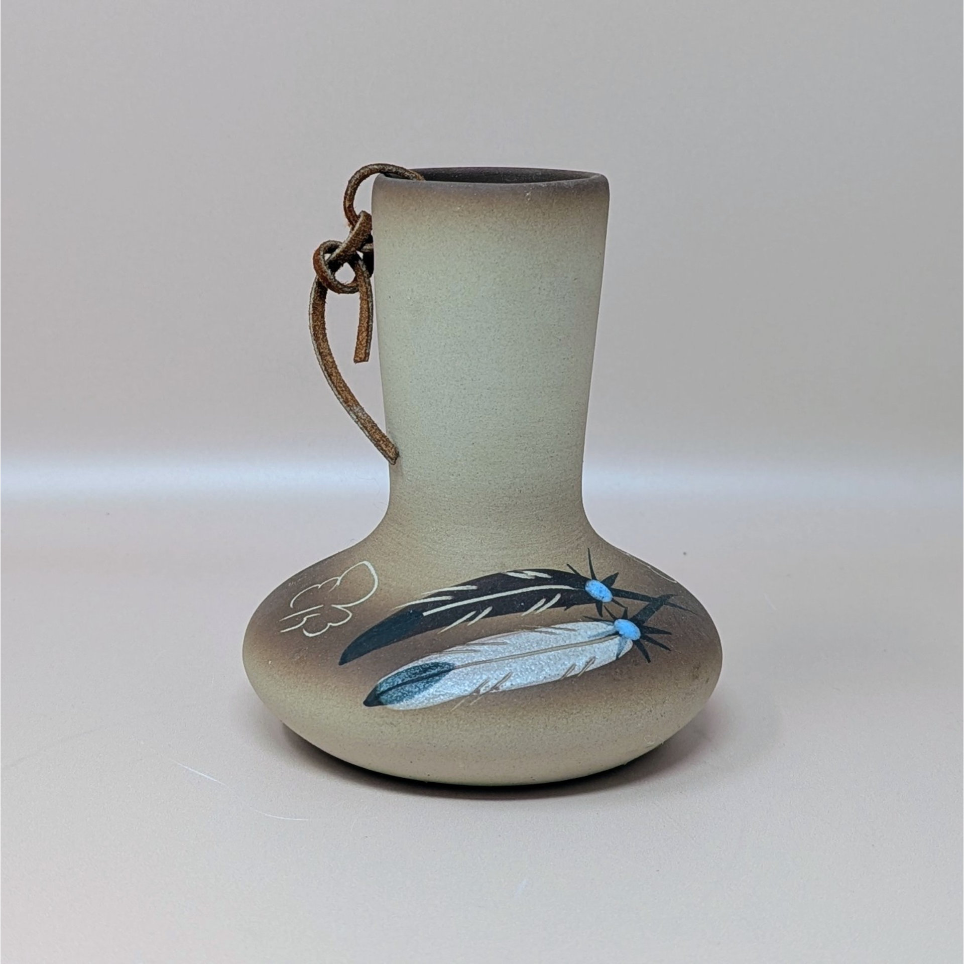 A hand painted tan pottery vase by Betty Selby featuring black and white feather motifs with turquoise accents and a leather strap tied around the neck.