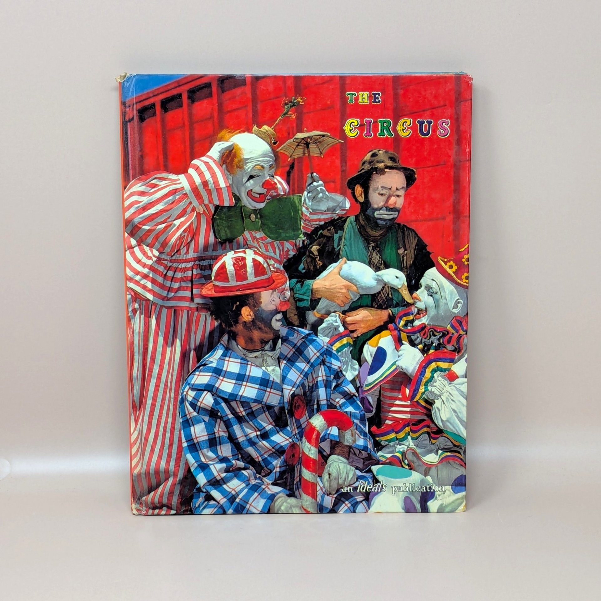 A 1961 vintage Ideals Publication book titled The Circus on a Dune colored background. The hardcover front features a vibrant red illustration of mid-century circus clowns in colorful striped and plaid costumes gathered together with a white duck.