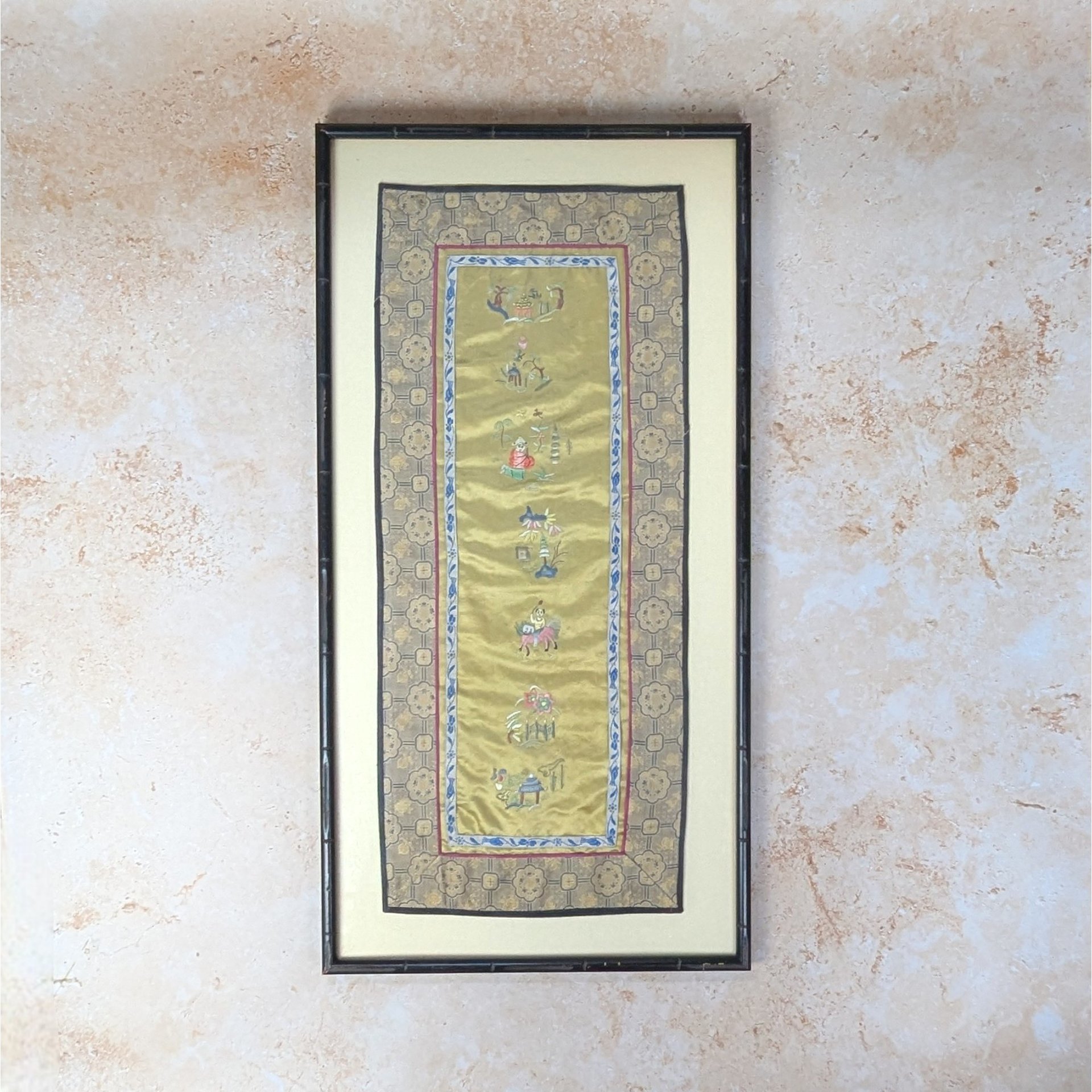 An antique Chinese silk embroidery sleeve band framed wall hanging on a tan textured background. The long vertical gold silk panel features intricate hand stitched figures and symbols inside a decorative patterned textile border and a dark wood faux bamboo frame.