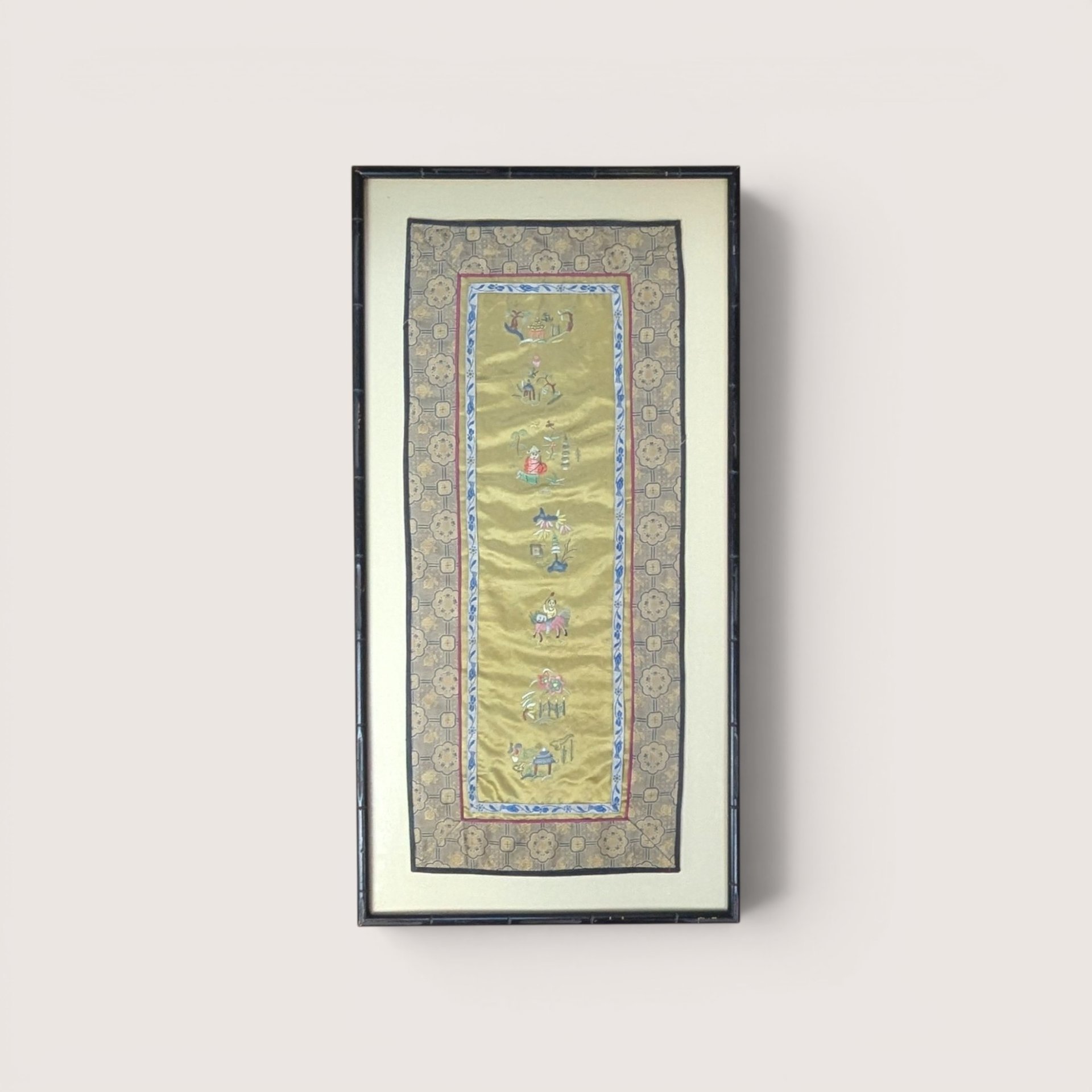 Framed vertical Chinese silk embroidery panel with intricate needlework figurines and a patterned border on a neutral wall