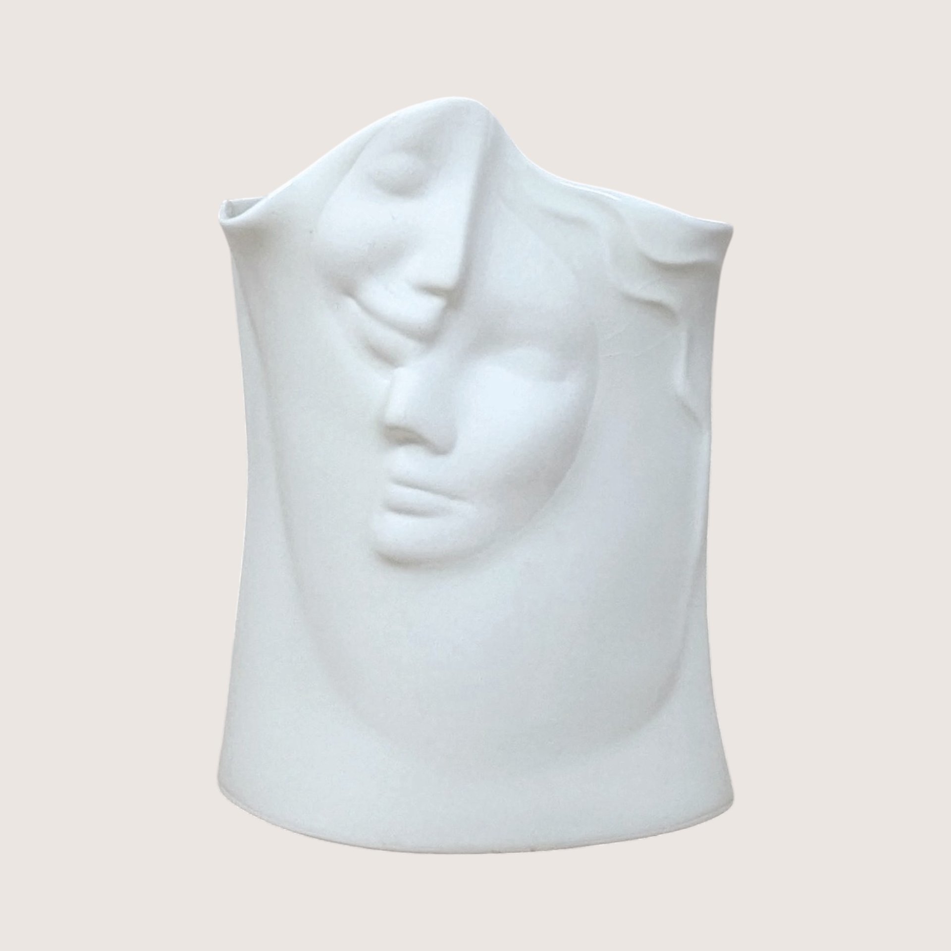 A matte white ceramic vase molded with two abstract interlocking faces, centered on a beige background.