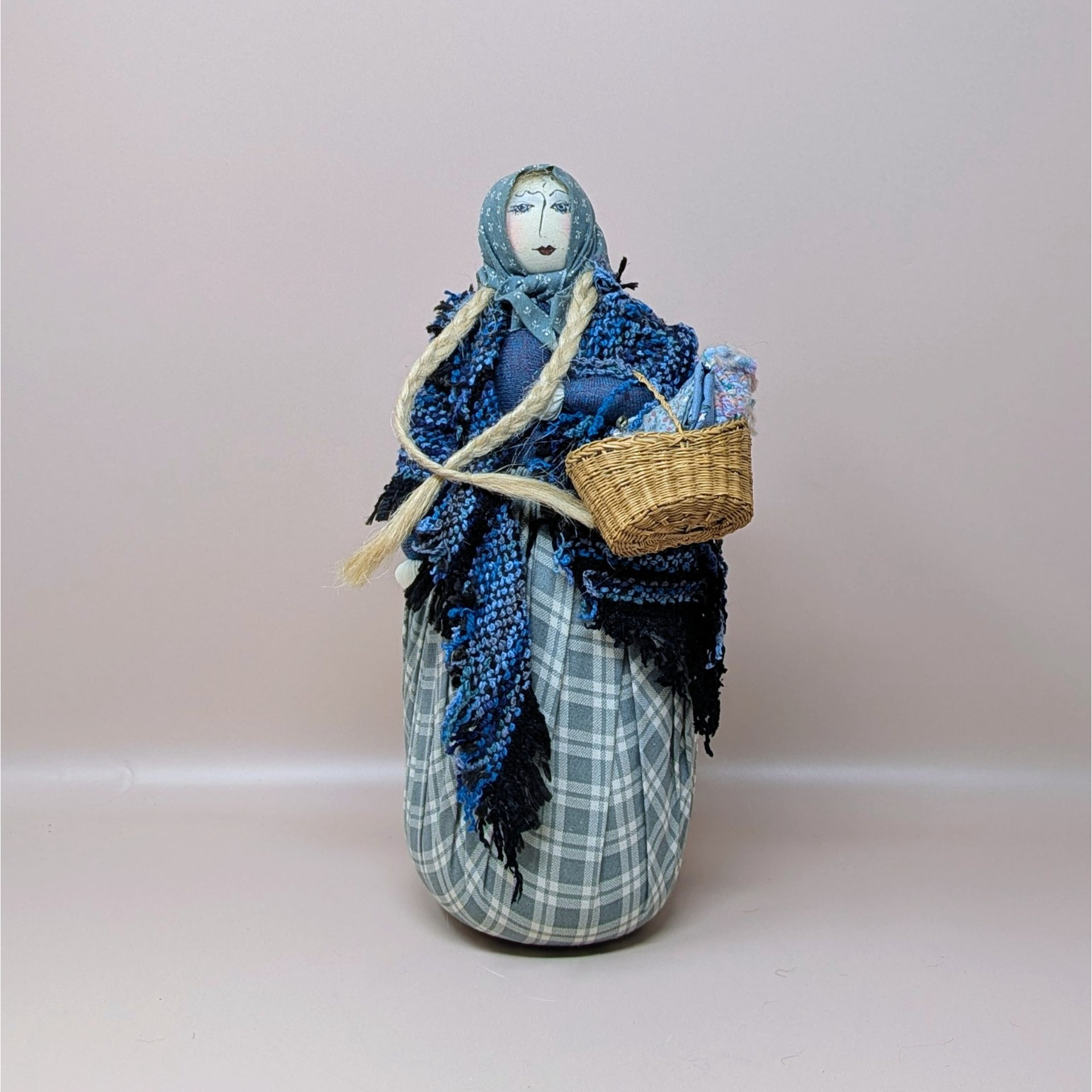 A detailed handmade folk art doll by Gretchen Lima featuring a woman in a blue shawl and plaid skirt. She has long flaxen braids, a blue headscarf, and holds a small woven basket, standing against a neutral background.
