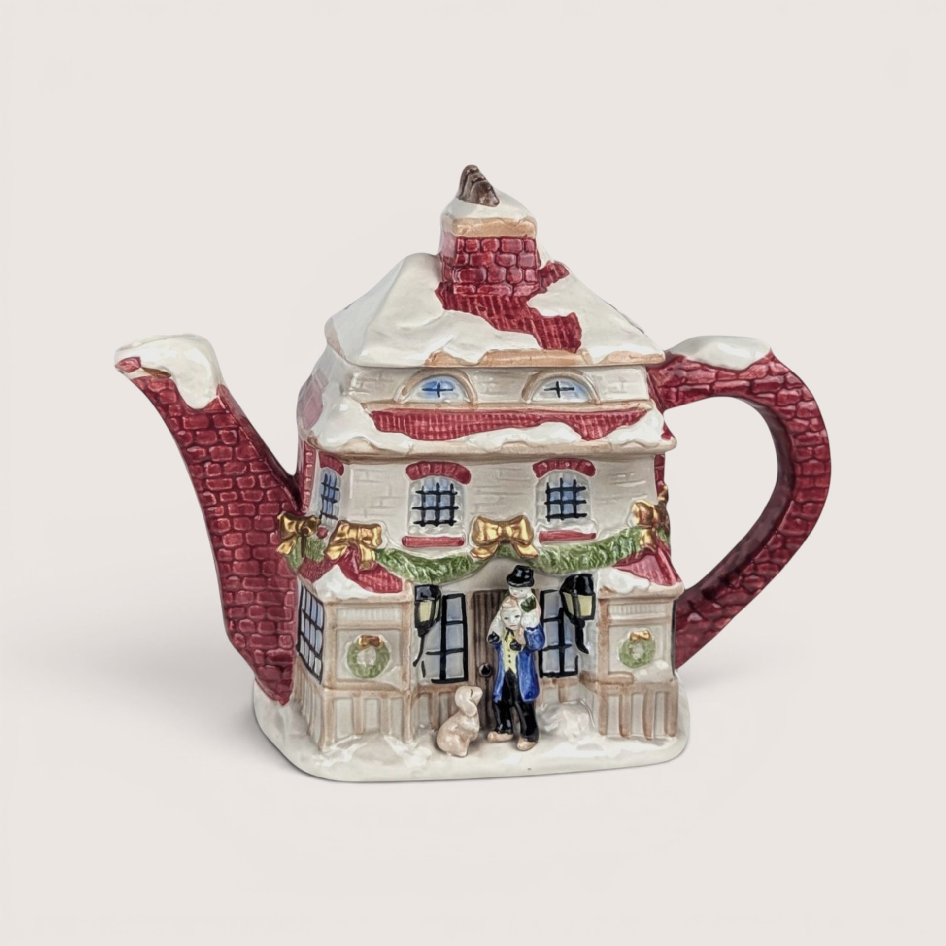 A ceramic teapot shaped like a snow-covered holiday house with festive garland, centered on a beige background.