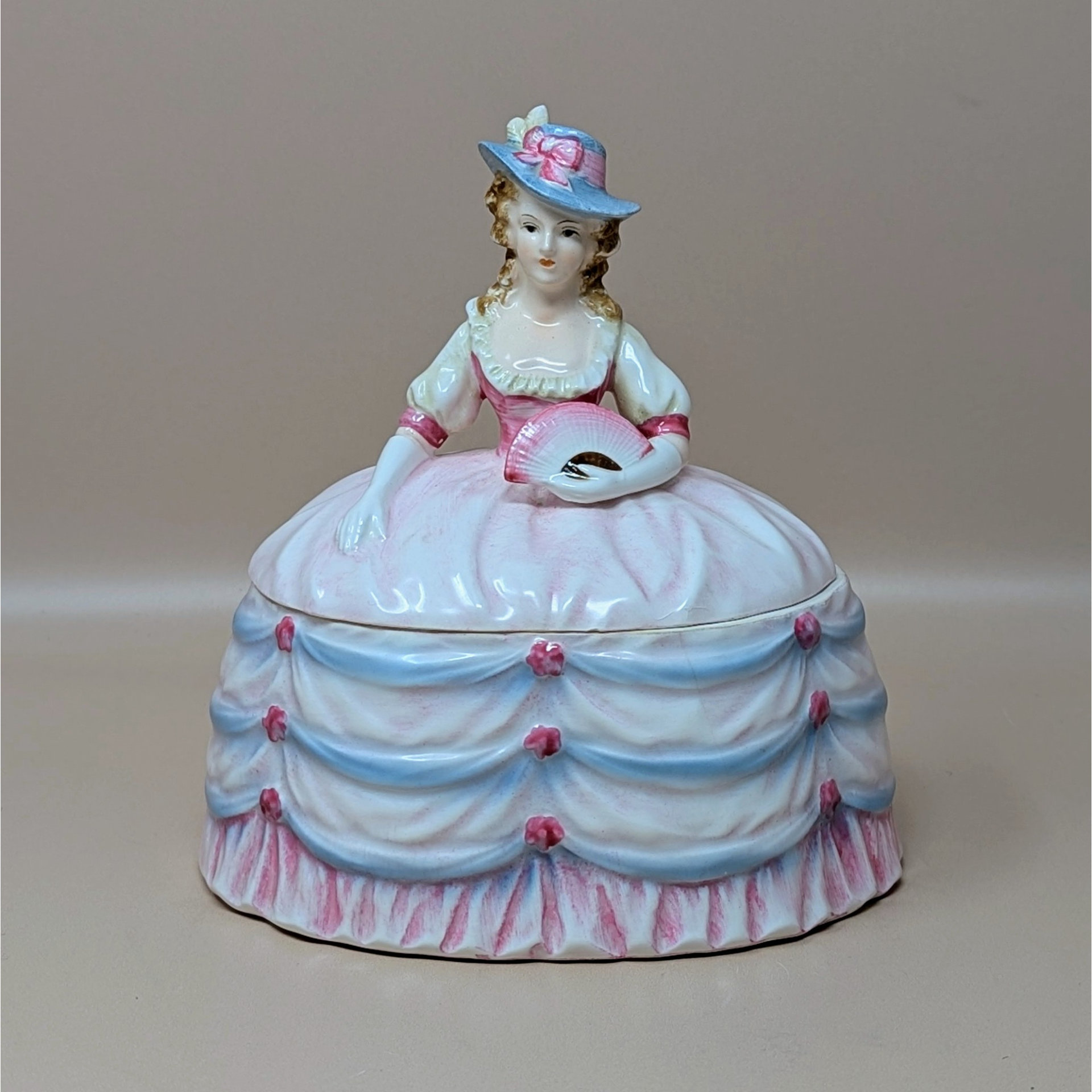 A vintage ceramic powder box shaped like a Victorian lady in a billowing pink and blue dress. She wears a wide-brimmed hat and holds a pink fan. The base of the skirt acts as the lid for the trinket dish.