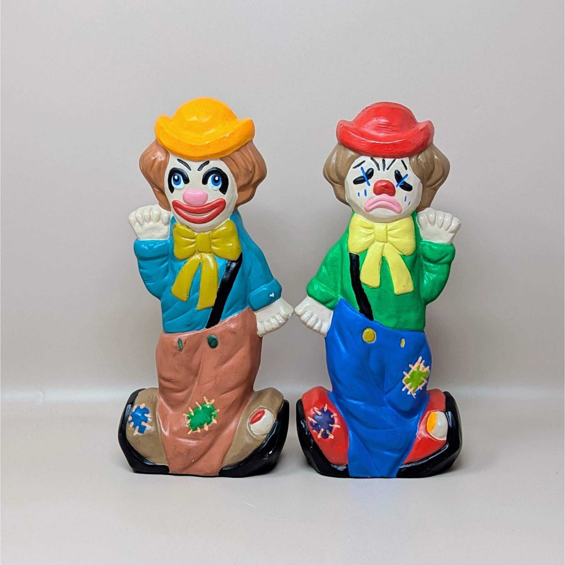 A pair of vintage ceramic clown banks. Each clown is double-sided, featuring a laughing face on one side and a crying face on the other. They wear colorful patched overalls and small hats.