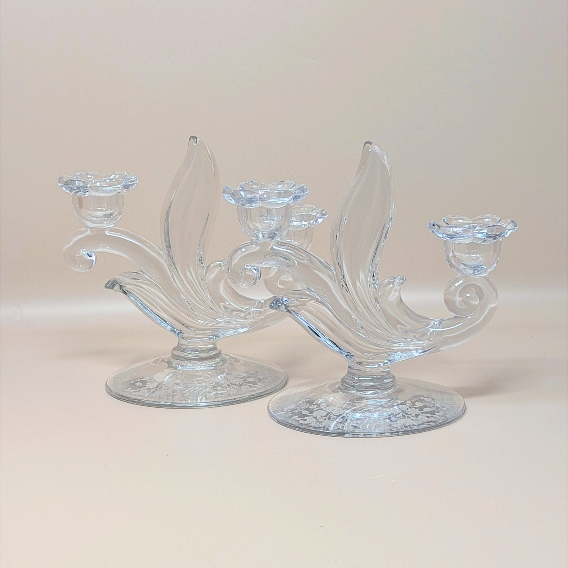 A pair of vintage clear glass double candlesticks by Duncan Miller in the First Love pattern. The holders feature elegant sweeping curves, flower-shaped candle cups, and an intricate etched floral design on the circular bases.