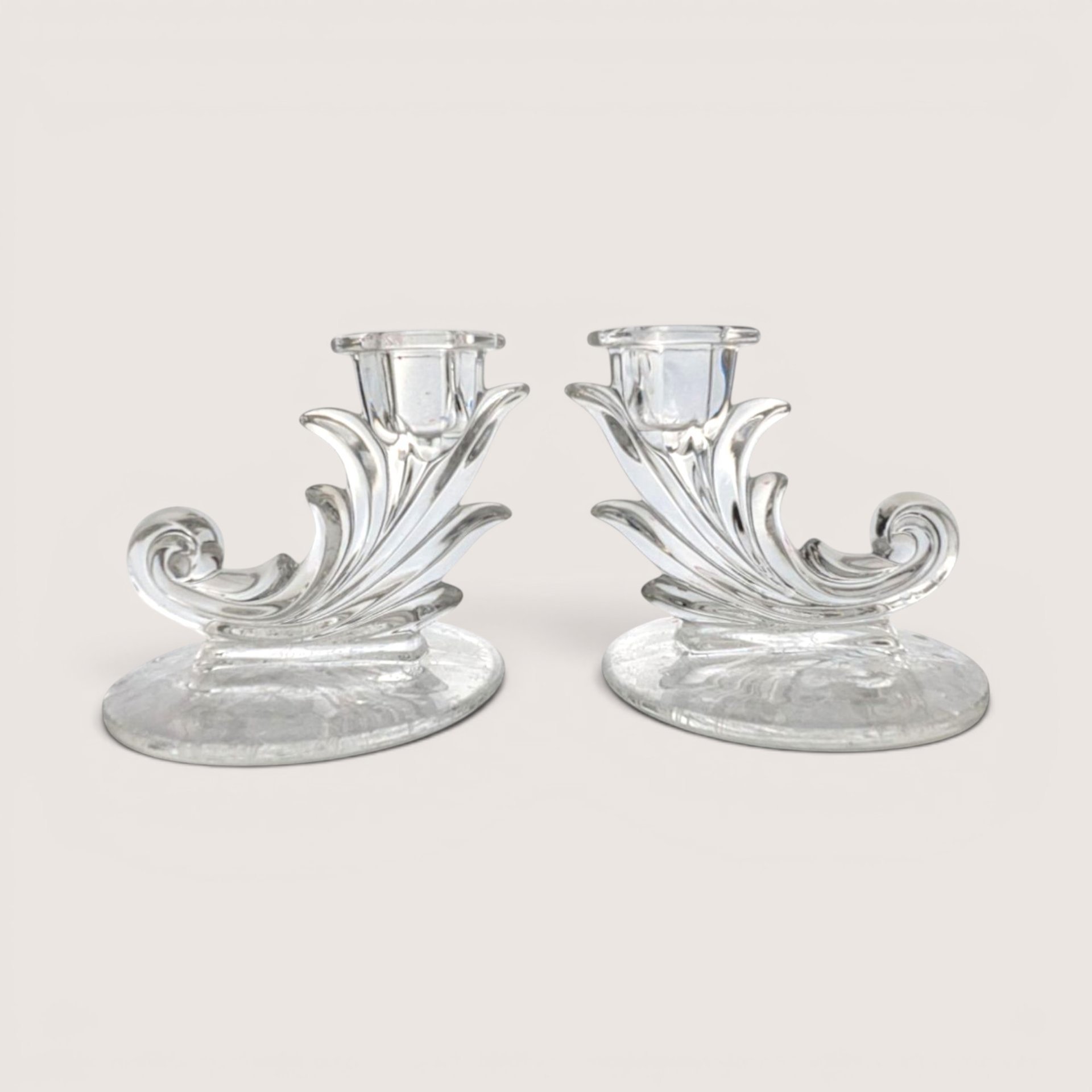 A pair of vintage clear glass cornucopia or leaf-shaped single candlestick holders, centered on a beige background.