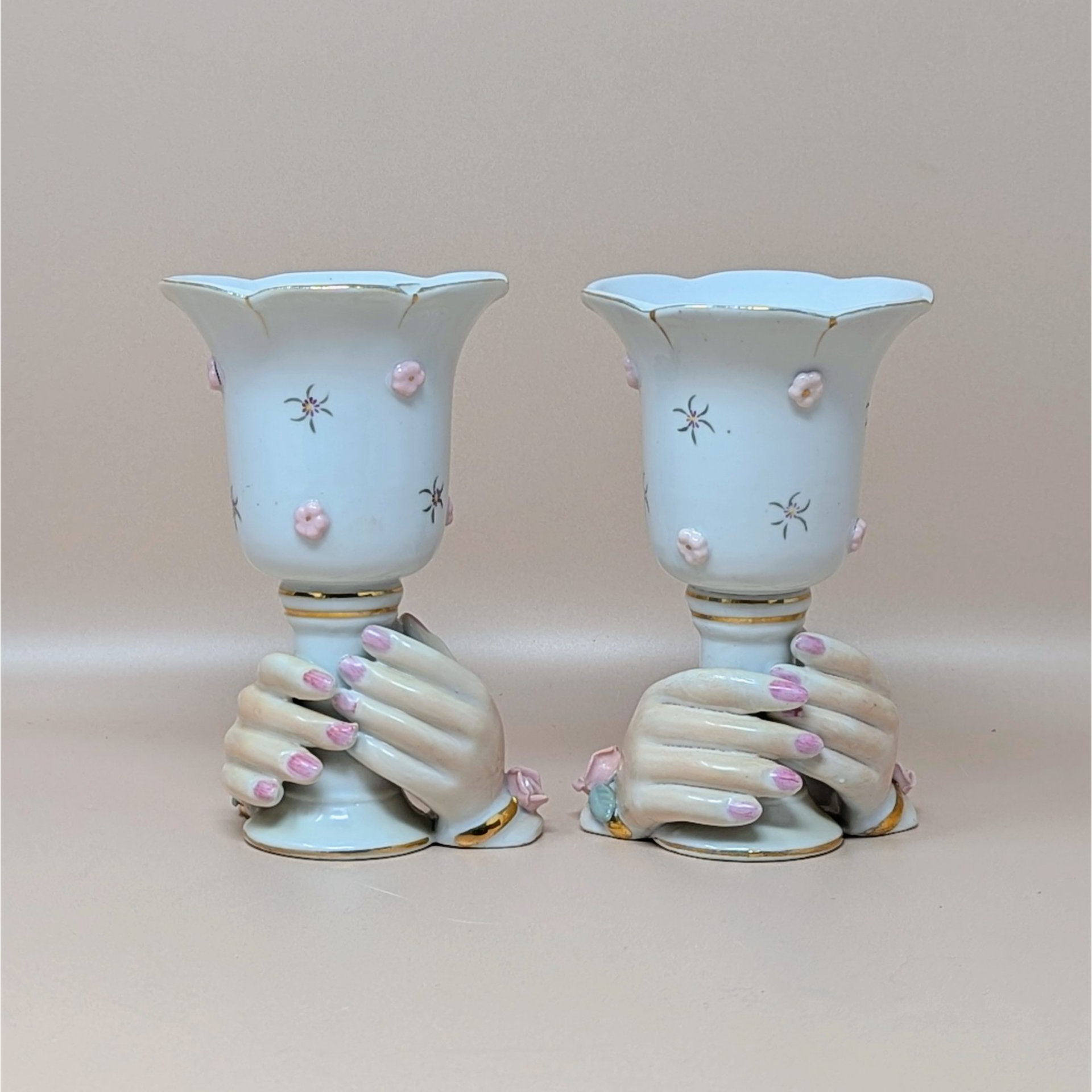 A pair of vintage ceramic candle holders featuring a pair of hands with pink fingernails clasping the base of a white cup. The cups are decorated with small pink 3D flowers and gold-rimmed scalloped edges.