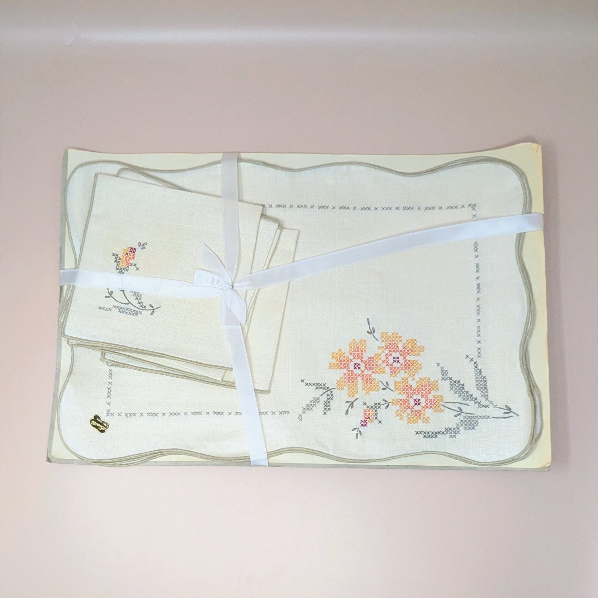 A vintage 8-piece Bucilla table linen set featuring four placemats and four matching napkins with colorful cross-stitch floral designs. The cream-colored linens are tied together with a white ribbon.