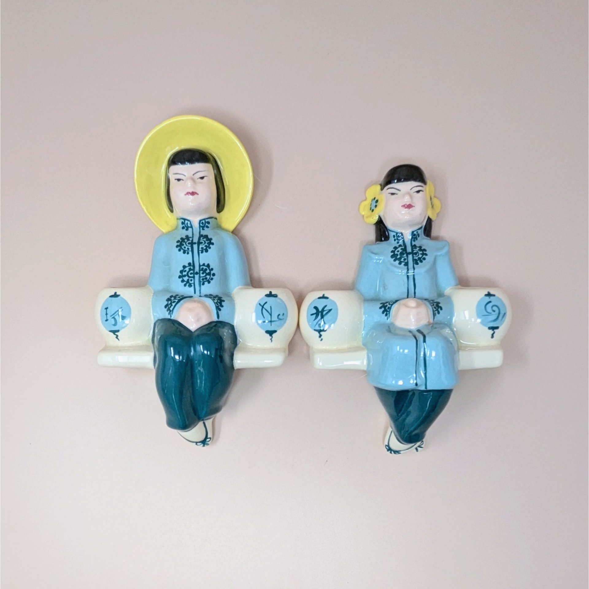 A pair of vintage Weil Ware ceramic wall-hanging candle holders. One depicts a man in a blue robe with a yellow hat, and the other depicts a woman in a blue robe with yellow flowers in her hair. Each figure sits between two small white lanterns that serve as candle holders.