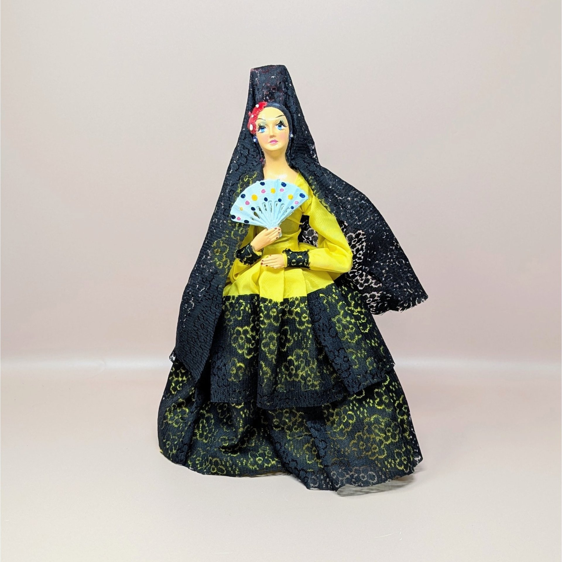 A vintage 12-inch doll depicting a Spanish senorita. She wears a bright yellow gown layered with intricate black lace, a matching black lace mantilla over her head, and holds a small painted fan.
