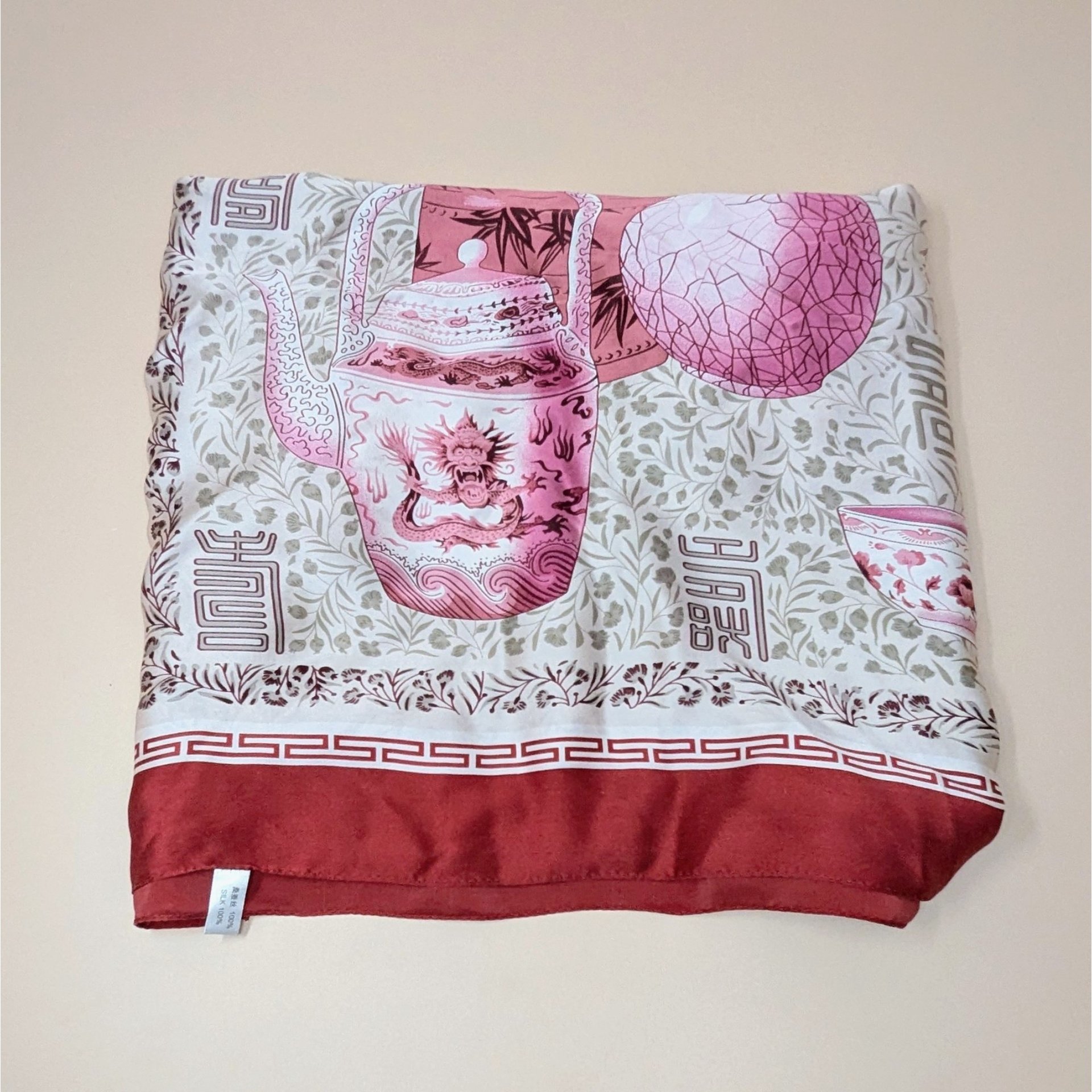 A vintage silk scarf featuring a large-scale print of a pink dragon-adorned teapot and ceramic bowls against a muted floral background. The scarf has a deep red border with a classic Greek key-style trim.