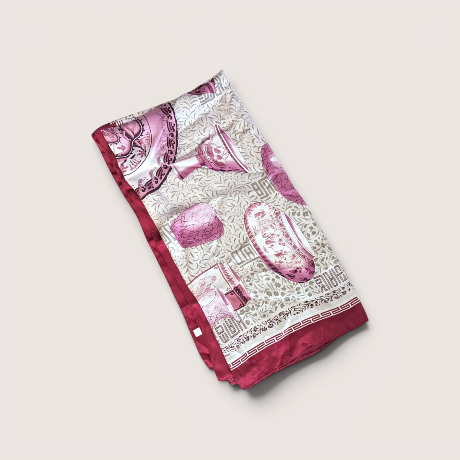 Folded silk scarf featuring pink Asian porcelain and pottery illustrations on a cream foliage pattern background with a dark red border, centered on a plain linen background.