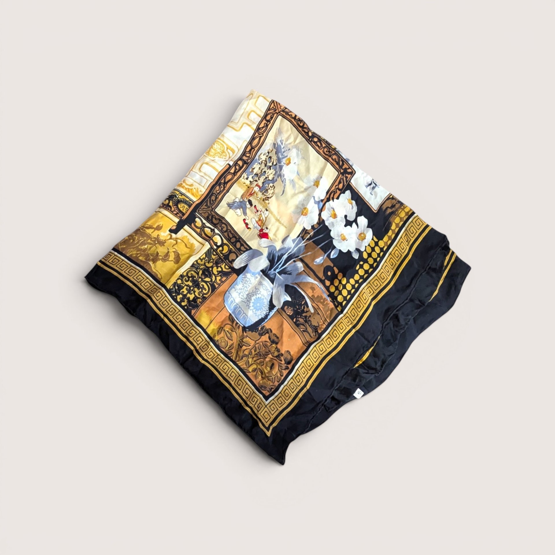 Folded scarf featuring a central blue and white vase with white flowers against an ornate gold, bronze, and black patterned background, centered on a plain linen background.