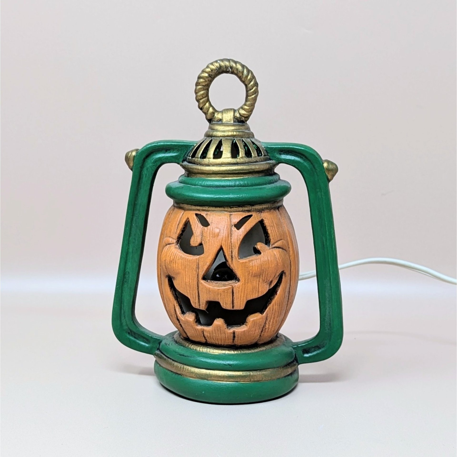 A vintage ceramic jack-o'-lantern light shaped like an old-fashioned lantern. The pumpkin is orange with a carved grin, set inside a green frame with gold detailing. An electrical cord is attached for illumination.