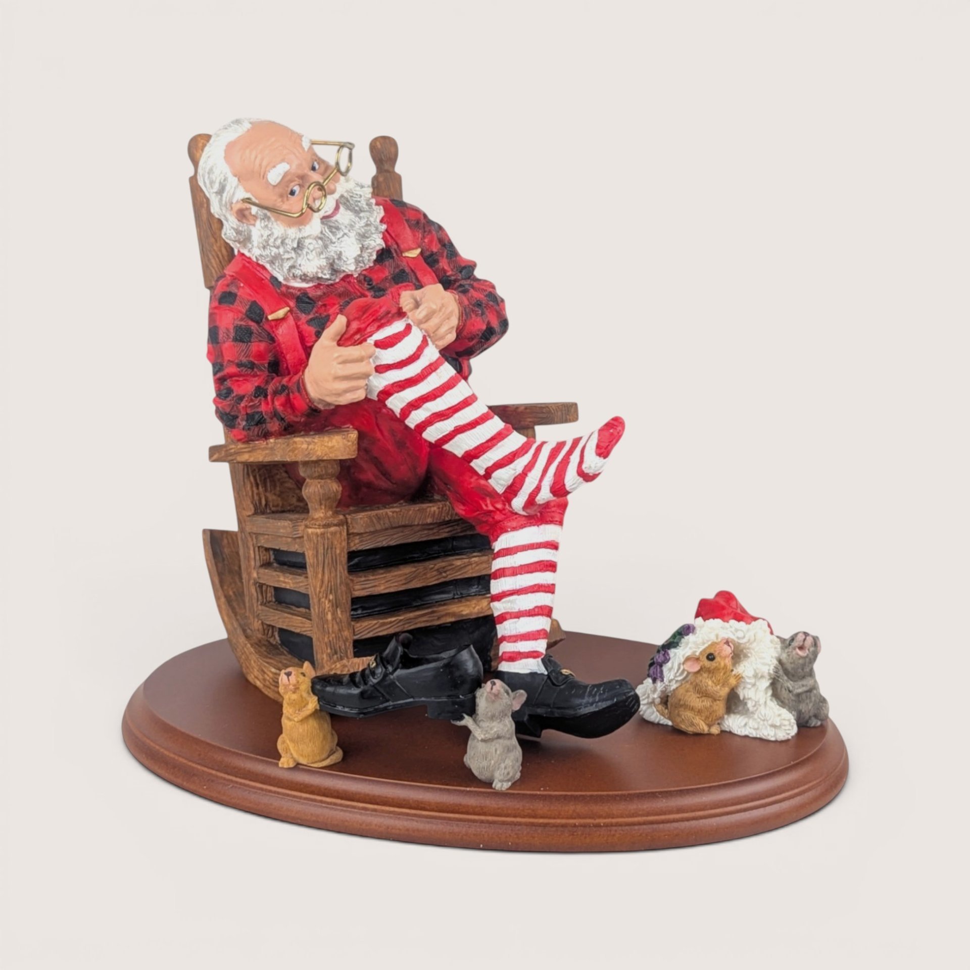 Santa Claus figurine sitting in a rocking chair mending a red and white striped stocking, surrounded by small mice on a wooden base, centered on a plain linen background.