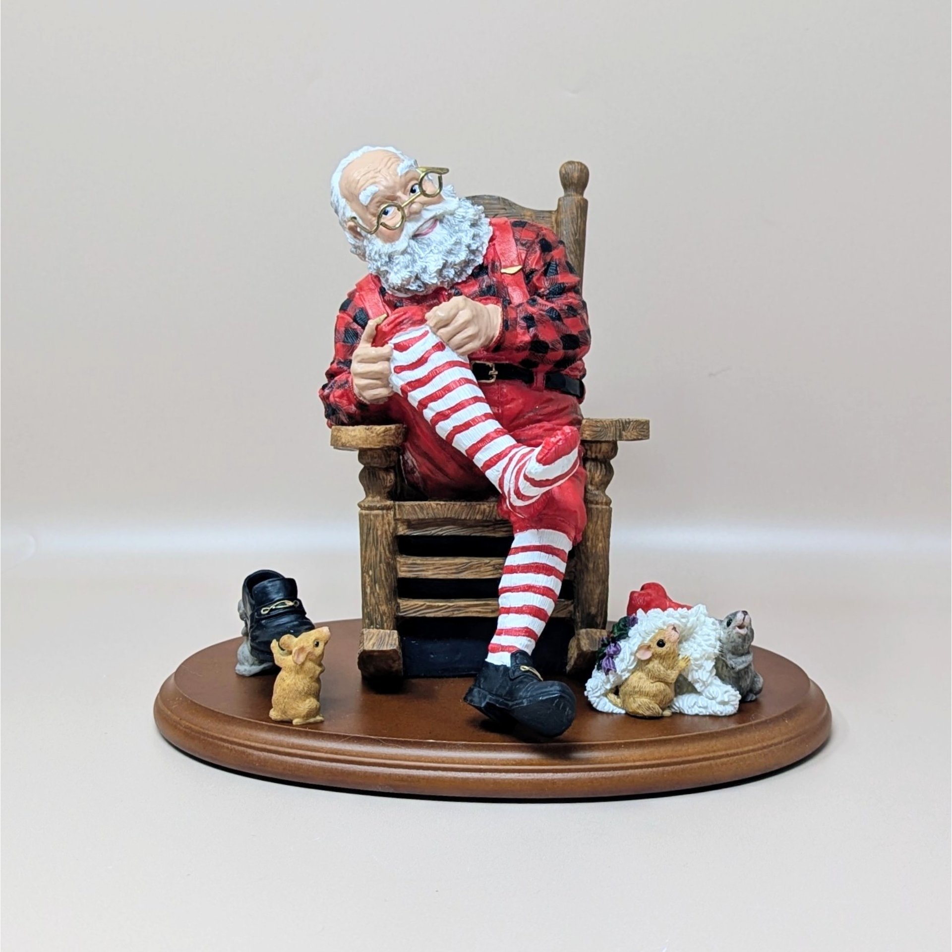 A vintage Department 56 Santa's Dress Rehearsal figure set. It features Santa in a red and black buffalo plaid shirt sitting in a wooden rocking chair, putting on a red-and-white striped stocking. Two small mice figures and a shoe are included on a wooden display base.