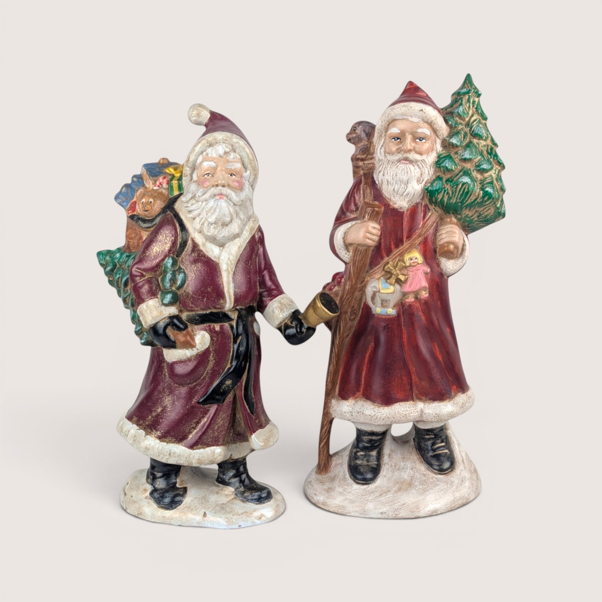 Pair of vintage Santa Claus figurines carrying Christmas trees and sacks of toys, centered on a plain linen background.
