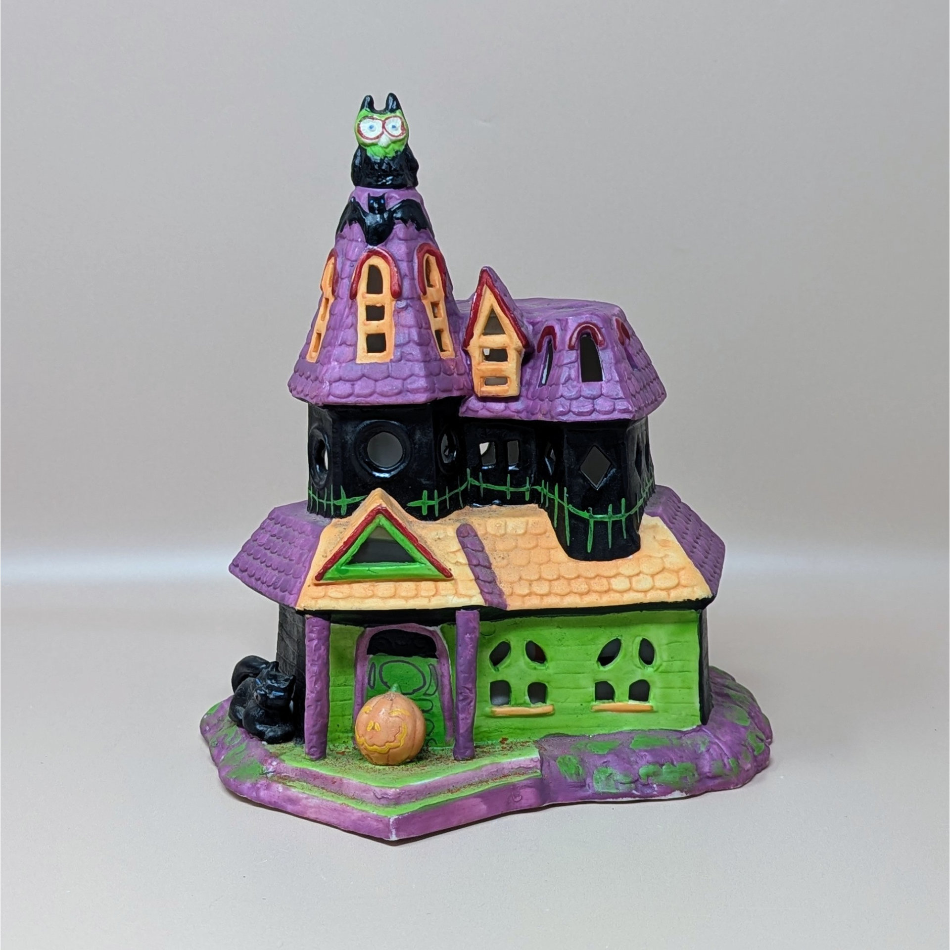 A vintage Celebrations by Silvestri ceramic Halloween house. It features a pointed purple roof, a bright green facade, and playful details like an owl on the tower, bats, and a small jack-o'-lantern at the front entrance.