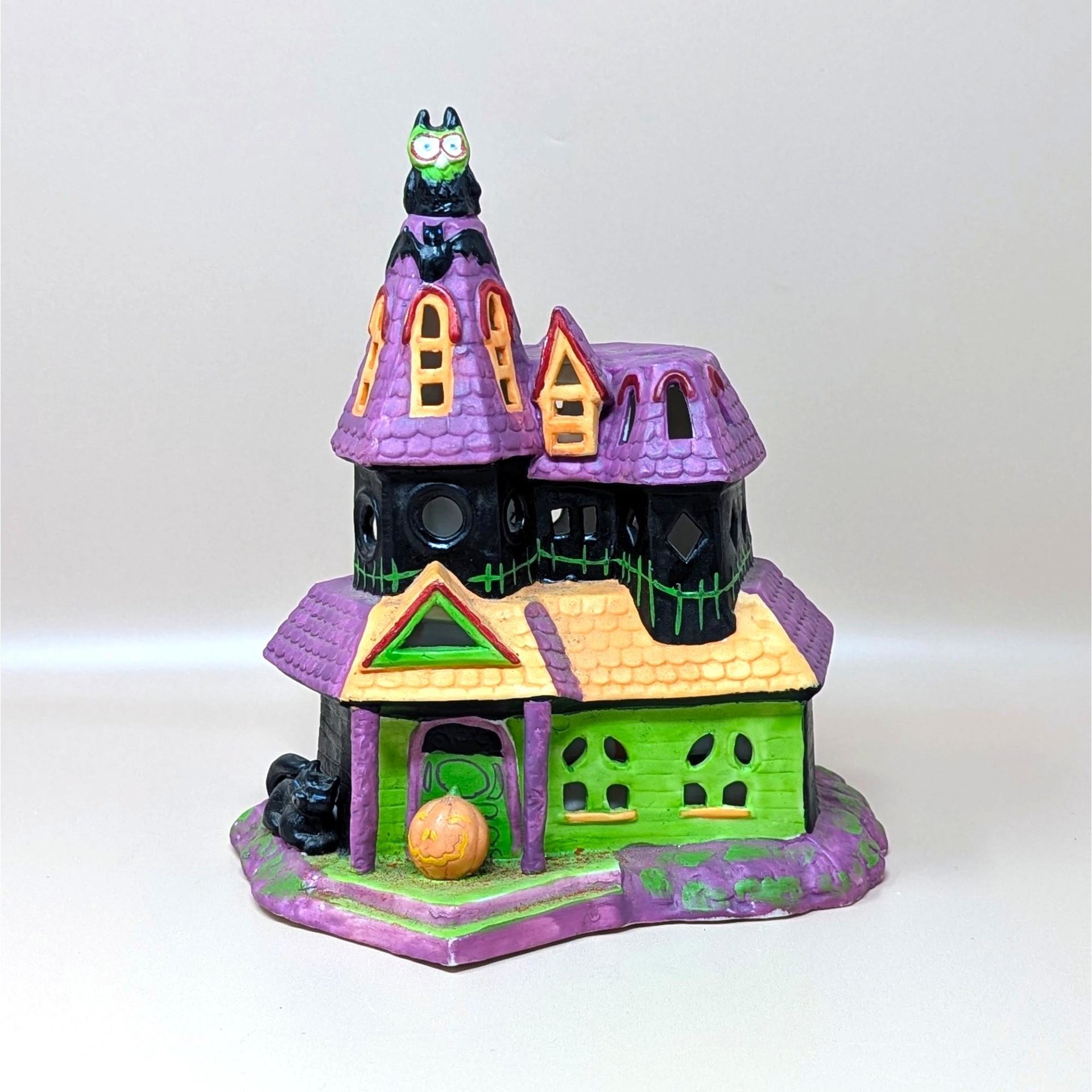 A vintage Celebrations by Silvestri ceramic Halloween house. It features a pointed purple roof, a bright green facade, and playful details like an owl on the tower, bats, and a small jack-o'-lantern at the front entrance.