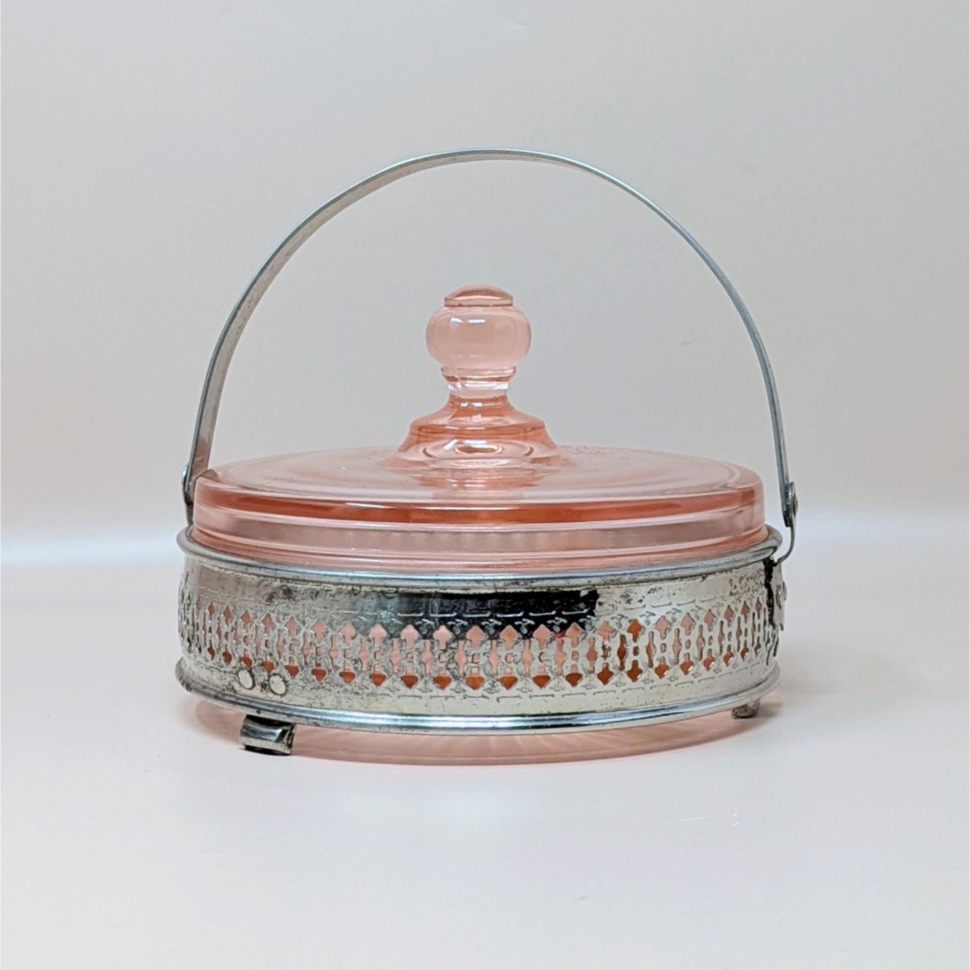 A vintage pink depression glass divided bowl resting in an ornate, silver-tone metal basket with a hinged handle. The matching pink glass lid features a round knob finial.