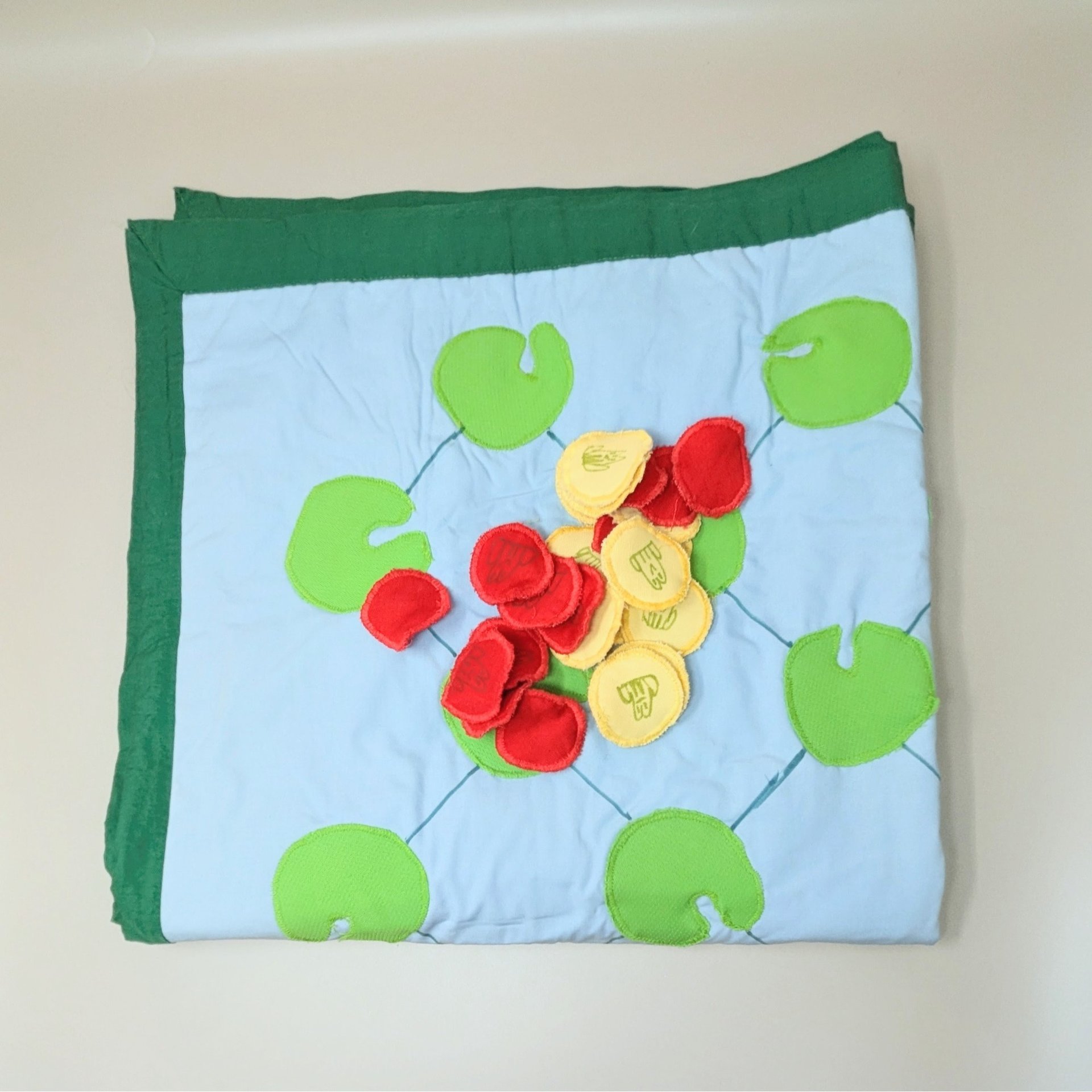 A handmade fabric game board featuring light blue cotton with green lily pad appliqués. It includes a collection of 24 red and yellow felt frog markers for gameplay.