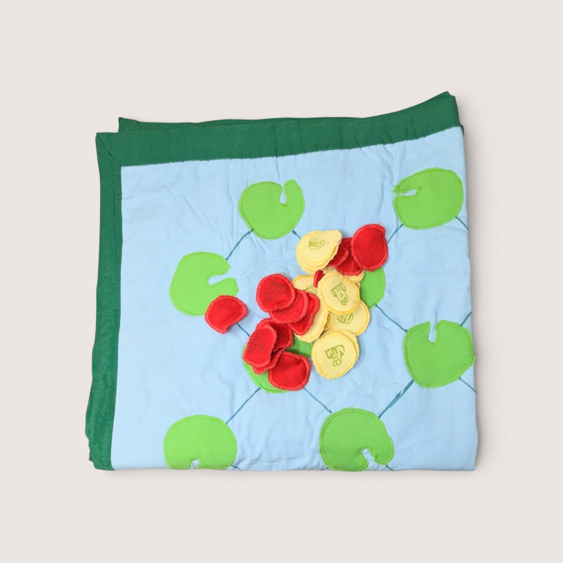 Folded fabric game mat featuring a light blue background patterned with green lily pads, topped with a pile of small red and yellow fabric tokens, centered on a plain linen background.