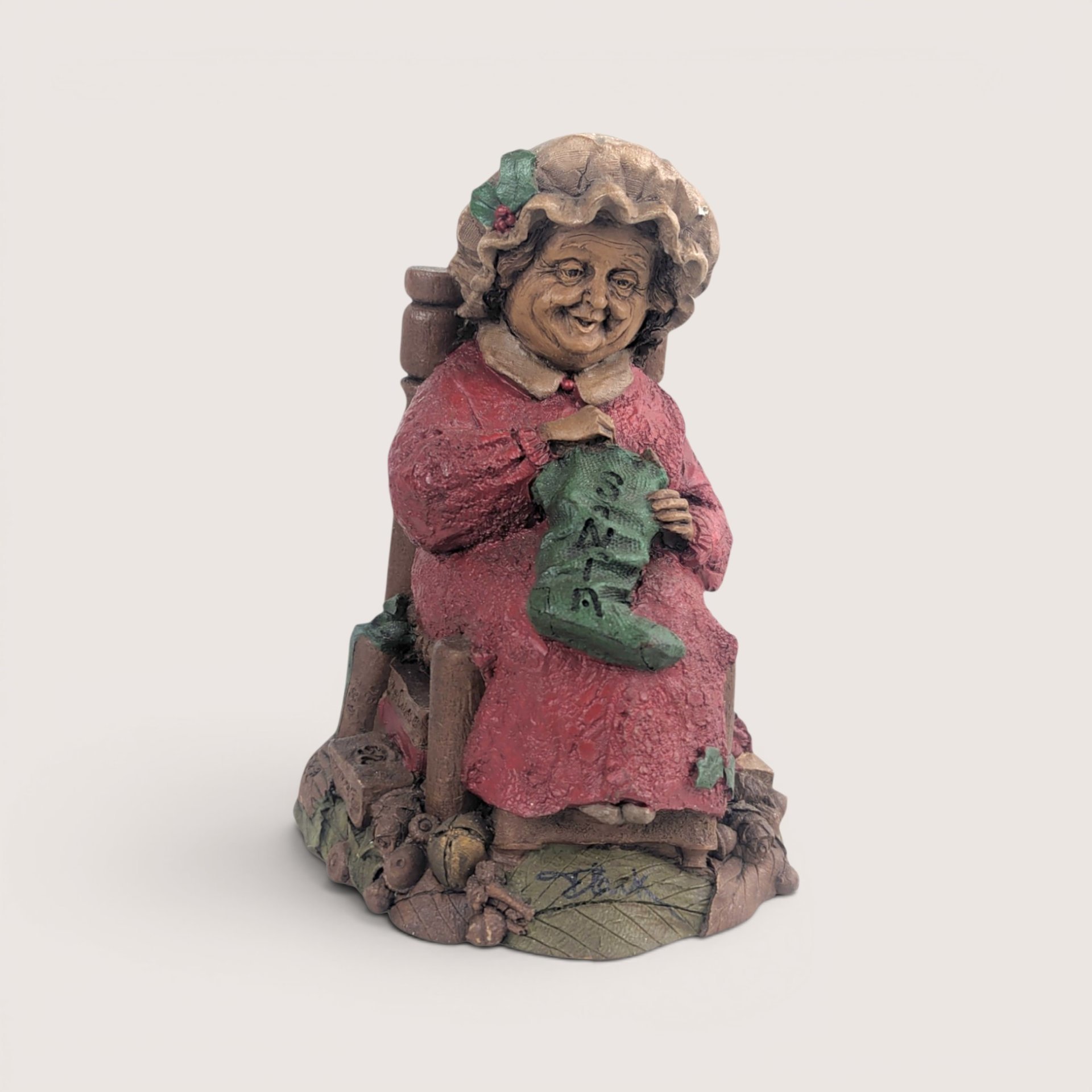 A vintage Tom Clark Cairn Studios resin figurine titled Mrs Claus featuring a woman sitting in a chair knitting a stocking.
