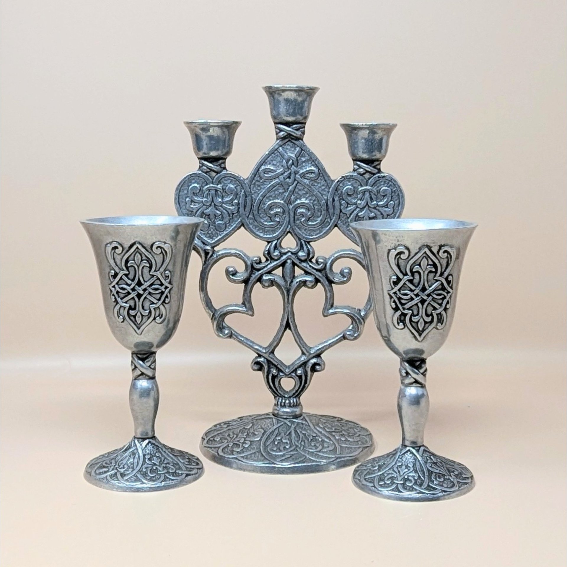 A 1996 Carlson Forevermore pewter set featuring a three-candle candelabra and two goblets, all decorated with ornate embossed knotwork and heart designs on a neutral backdrop.