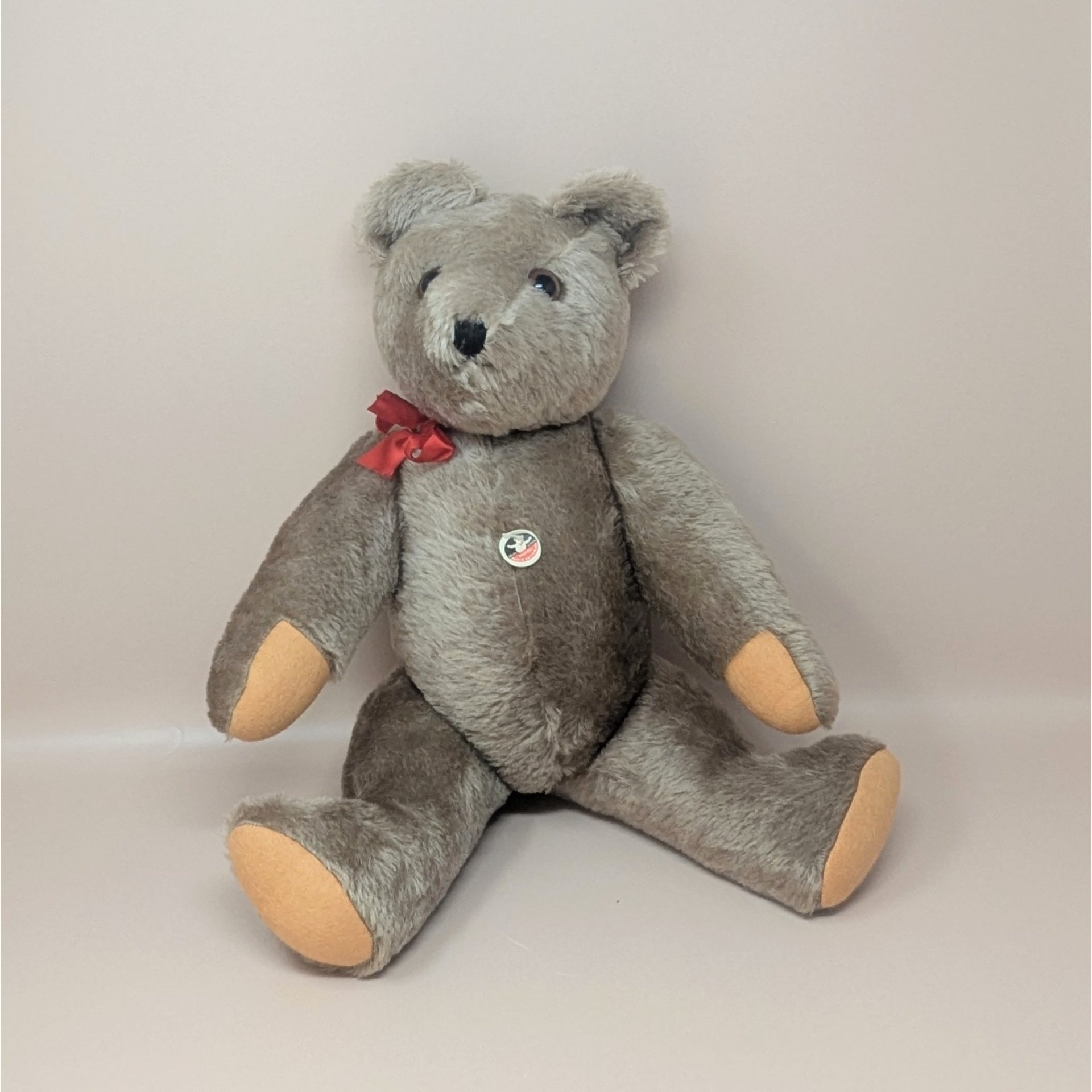 A vintage handmade Felpa teddy bear from Switzerland with grey-brown fur, a red bow, and tan paw pads, shown in a sitting position.