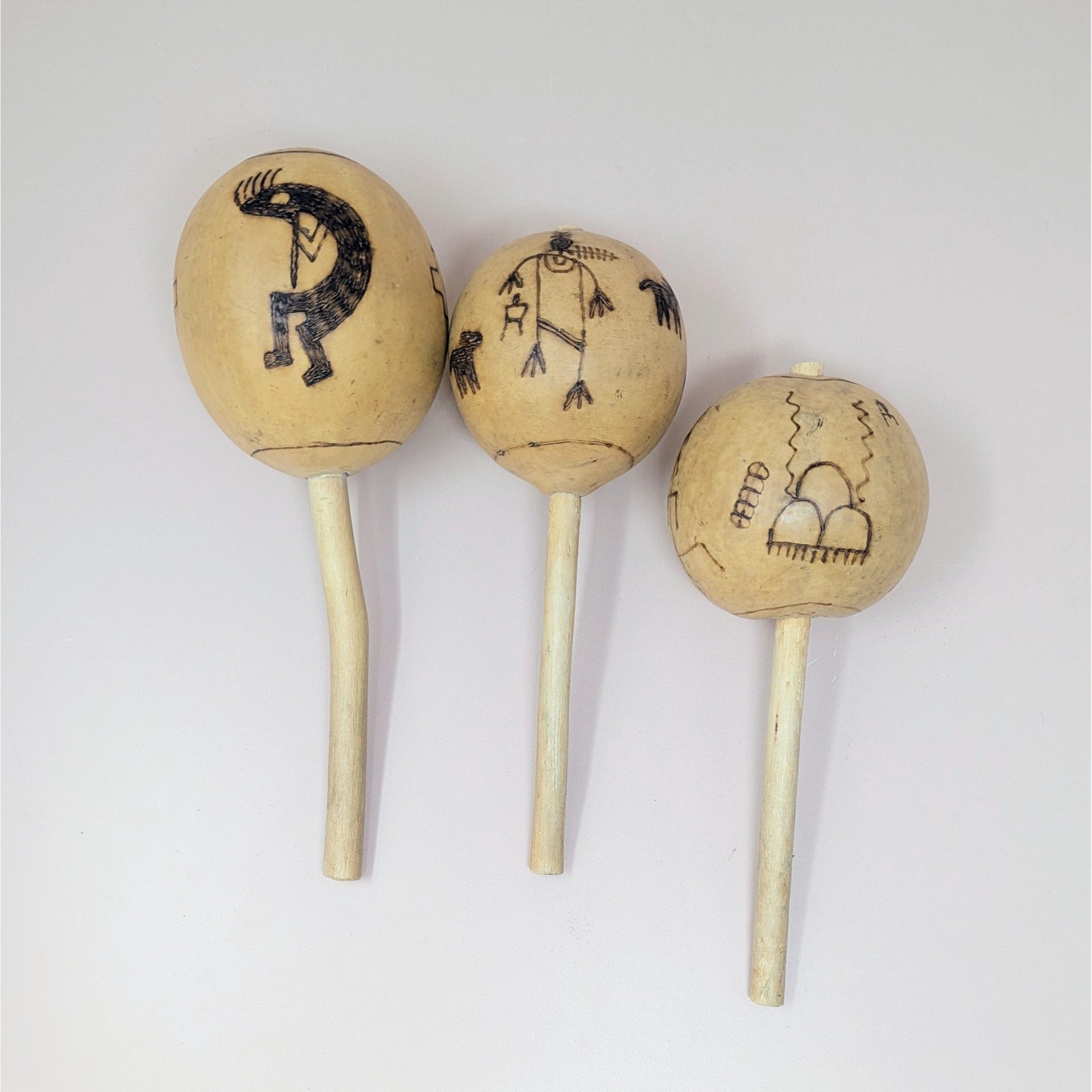 A trio of vintage hand-etched gourd maracas featuring Kokopelli and other traditional figures on simple wooden handles.