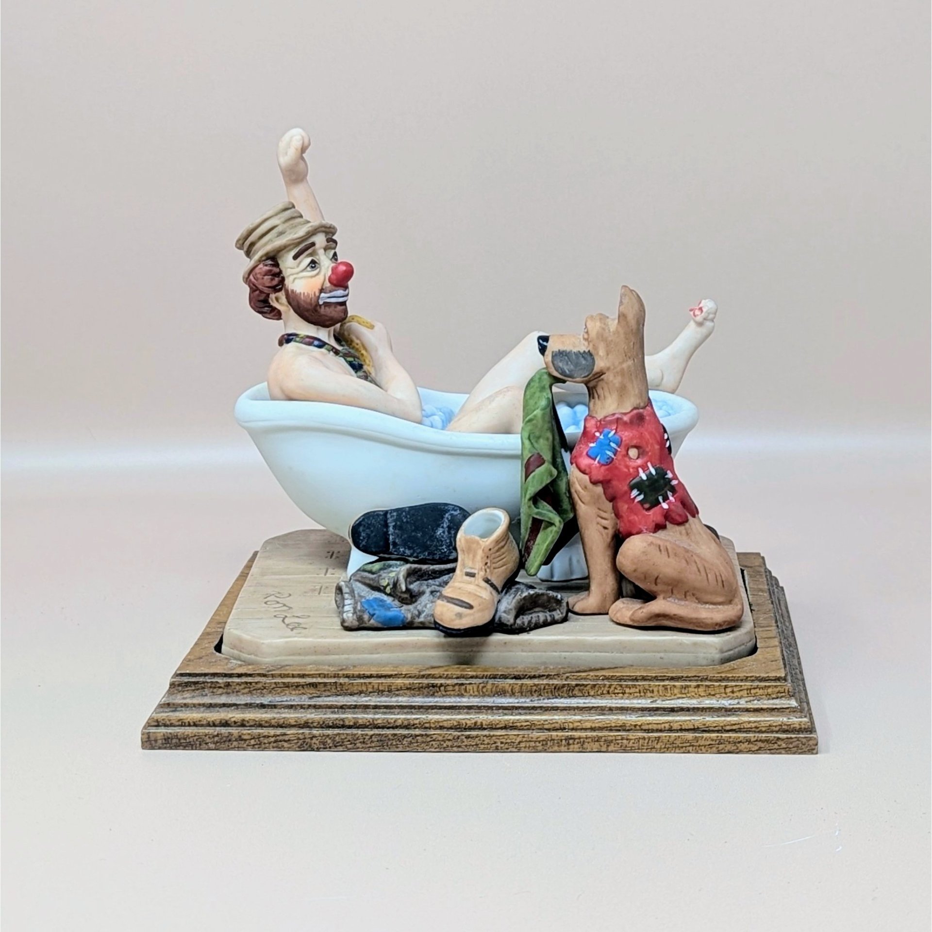 A vintage Ron Lee figurine titled A Hard Day's Bath featuring a clown in a bathtub with a dog holding a towel, mounted on a tiered wooden base.