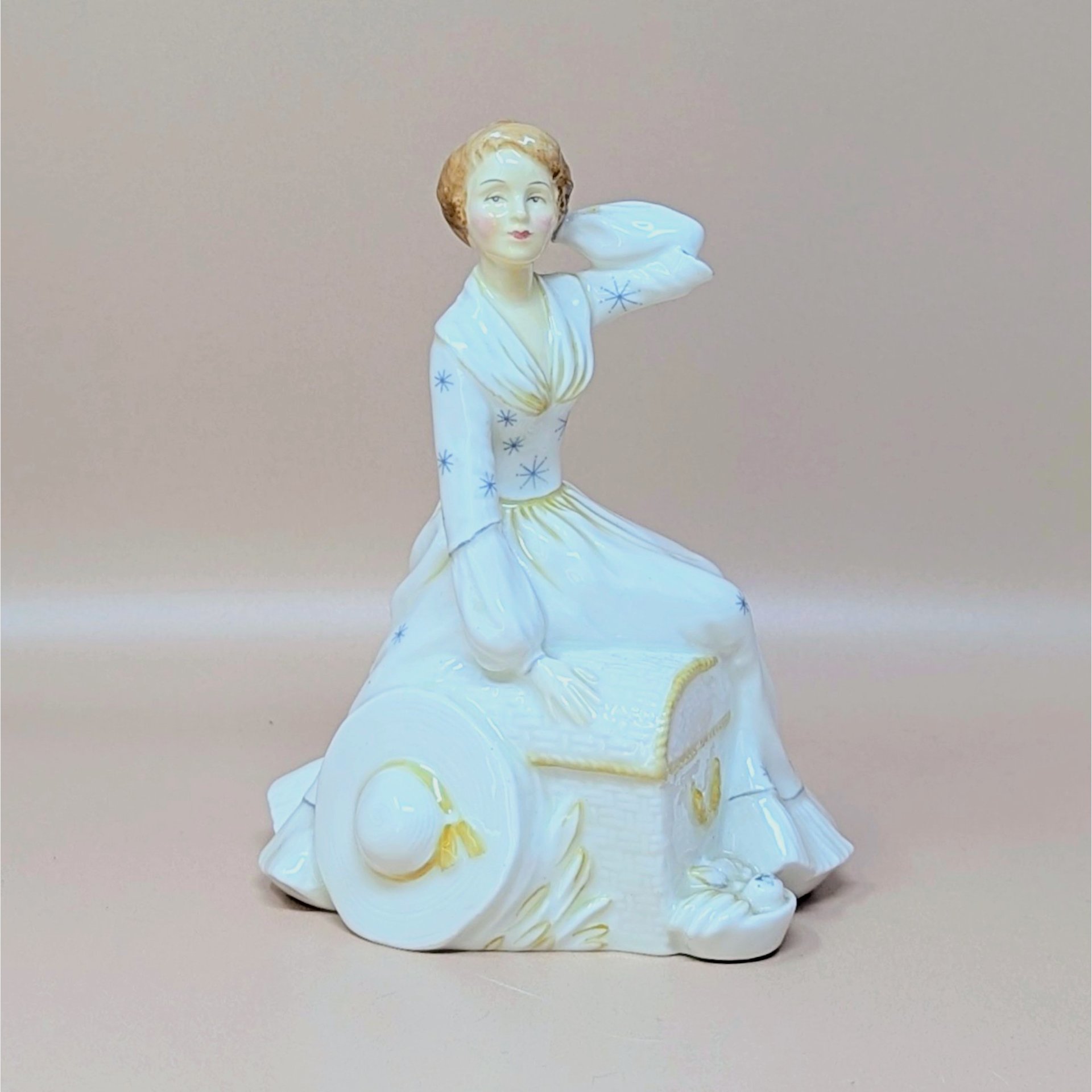A vintage Royal Doulton figurine titled Summers Day featuring a woman in a white dress with blue star patterns sitting next to a hat and basket