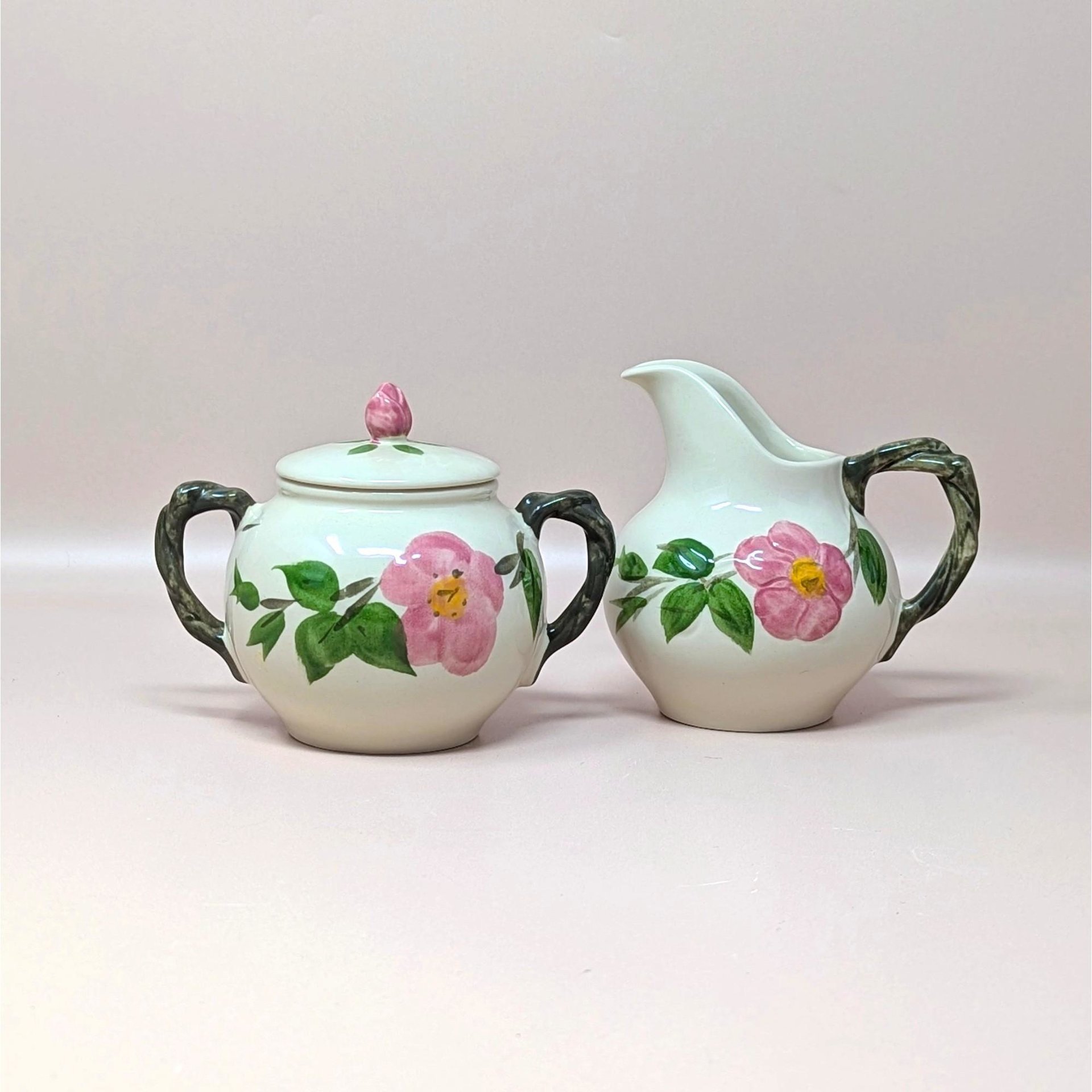 Hand painted Franciscan Desert Rose ceramic cream and sugar set on a soft pink background. The lidded sugar bowl and cream pitcher feature dimensional pink wild roses with green leaves and realistic brown branch handles and finial.