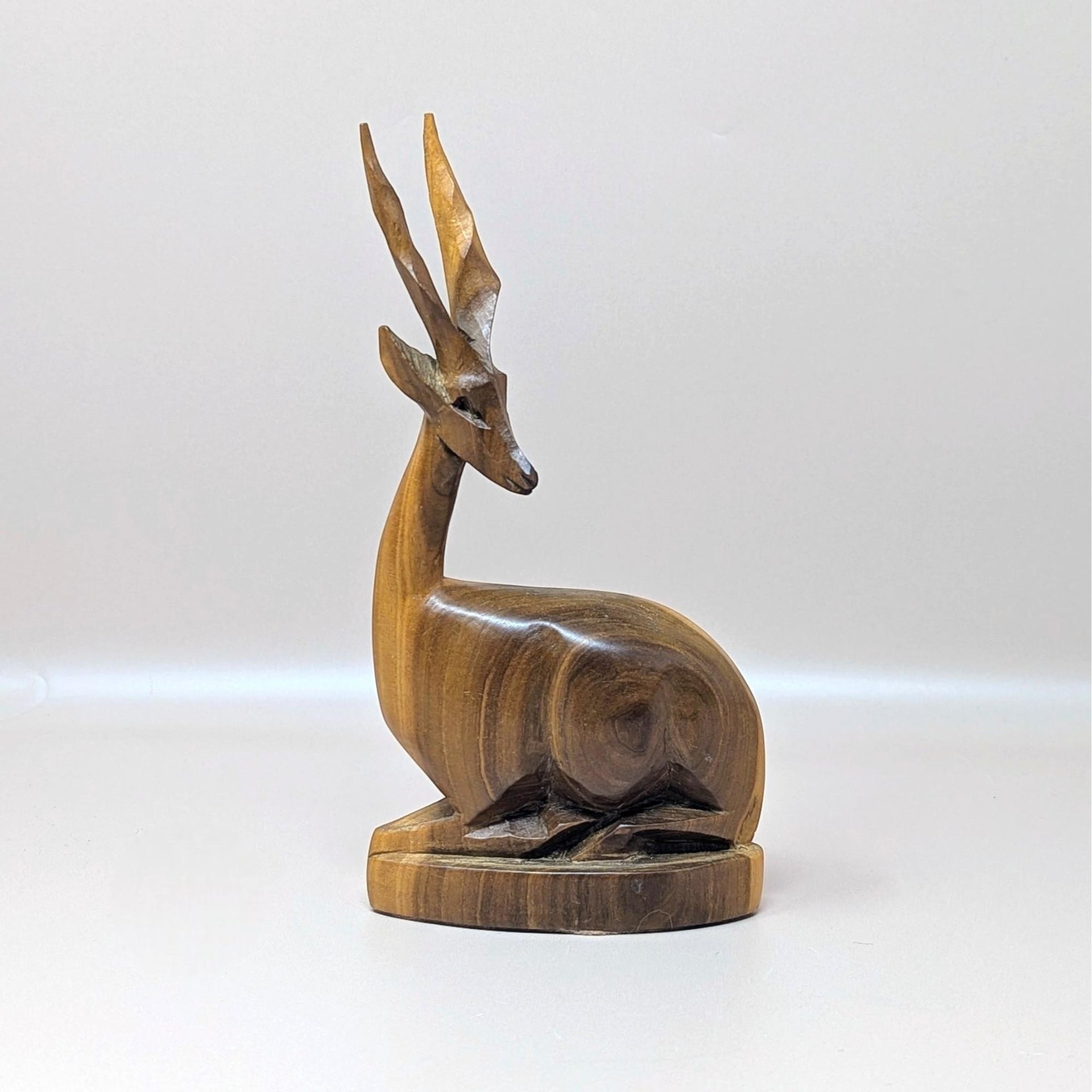 Hand carved vintage wooden gazelle figurine resting on a soft pink background. The sculpture depicts a stylized, seated antelope with long, twisted horns and a smooth, polished grain that highlights the rich brown tones of the wood.