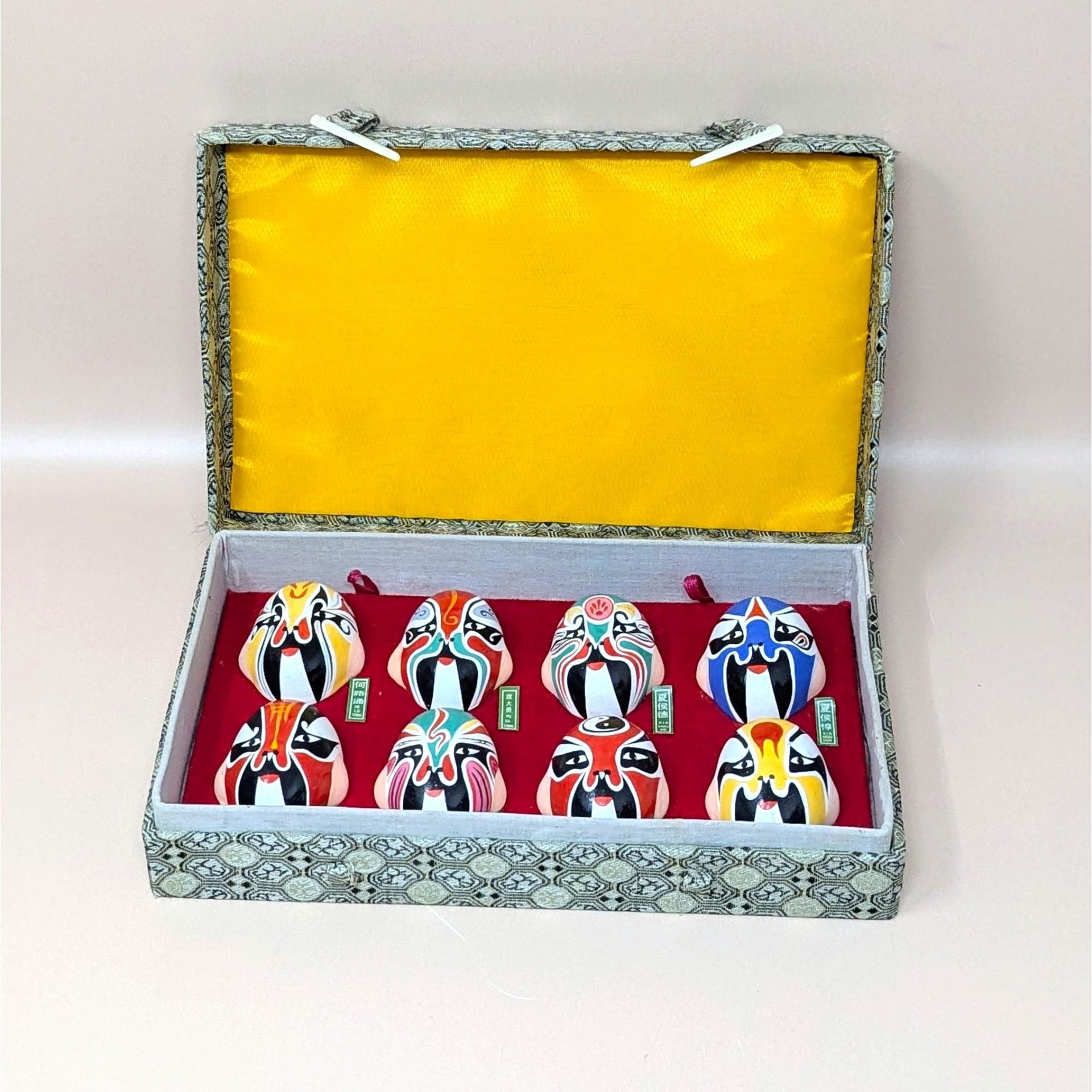 A vintage boxed display of eight hand painted Beijing Opera clay masks resting on a soft pink background. The vibrant, colorful theatrical facial makeup designs are arranged on red velvet inside a fabric covered case with a bright yellow satin lined lid.