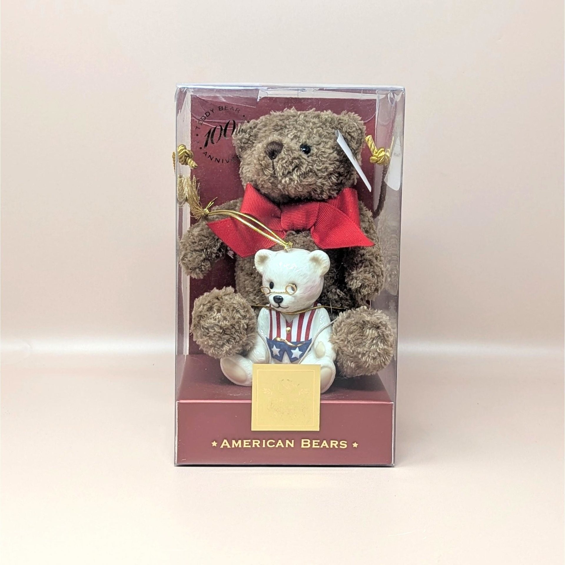 A sealed vintage Lenox American Bears 100th anniversary set on a soft pink background. The clear display box contains a brown plush teddy bear with a red bow and a smaller white porcelain bear ornament wearing a patriotic vest and tiny wire glasses.