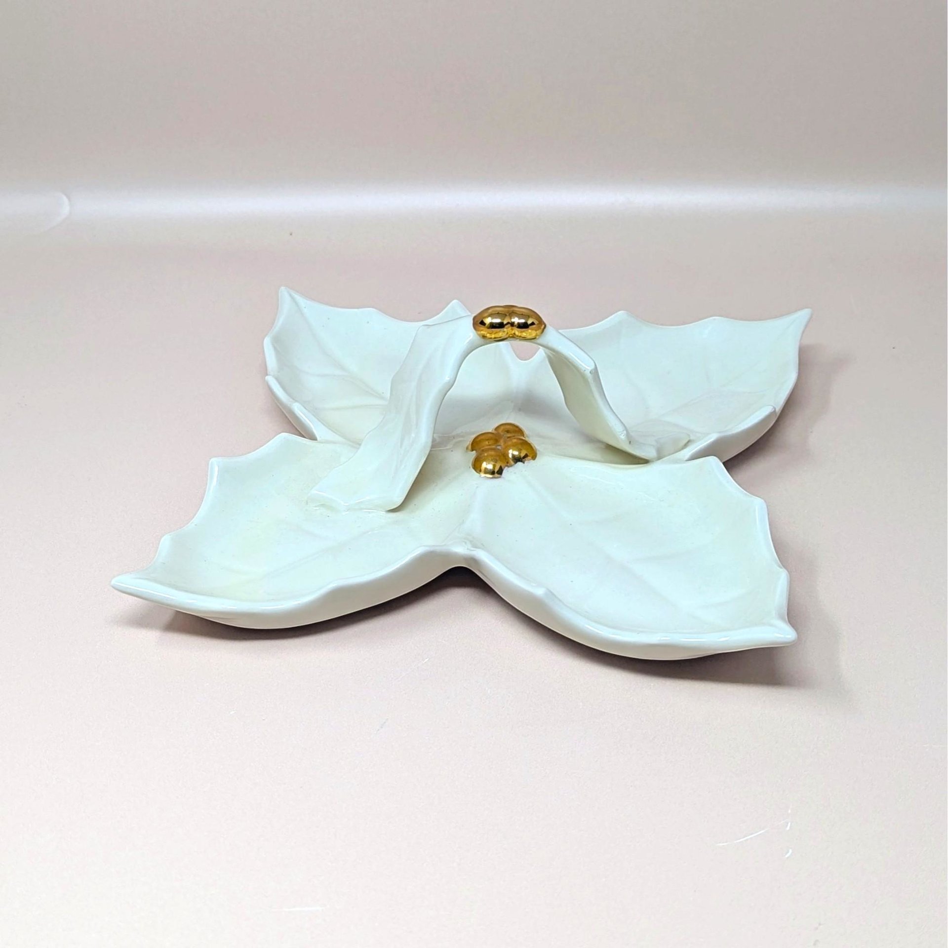 A vintage cream colored Holland Mold ceramic four part holly leaf dish resting on a soft pink background. The square hostess tray features a prominent central arched handle and hand painted shiny gold metallic berry clusters at the center and apex.