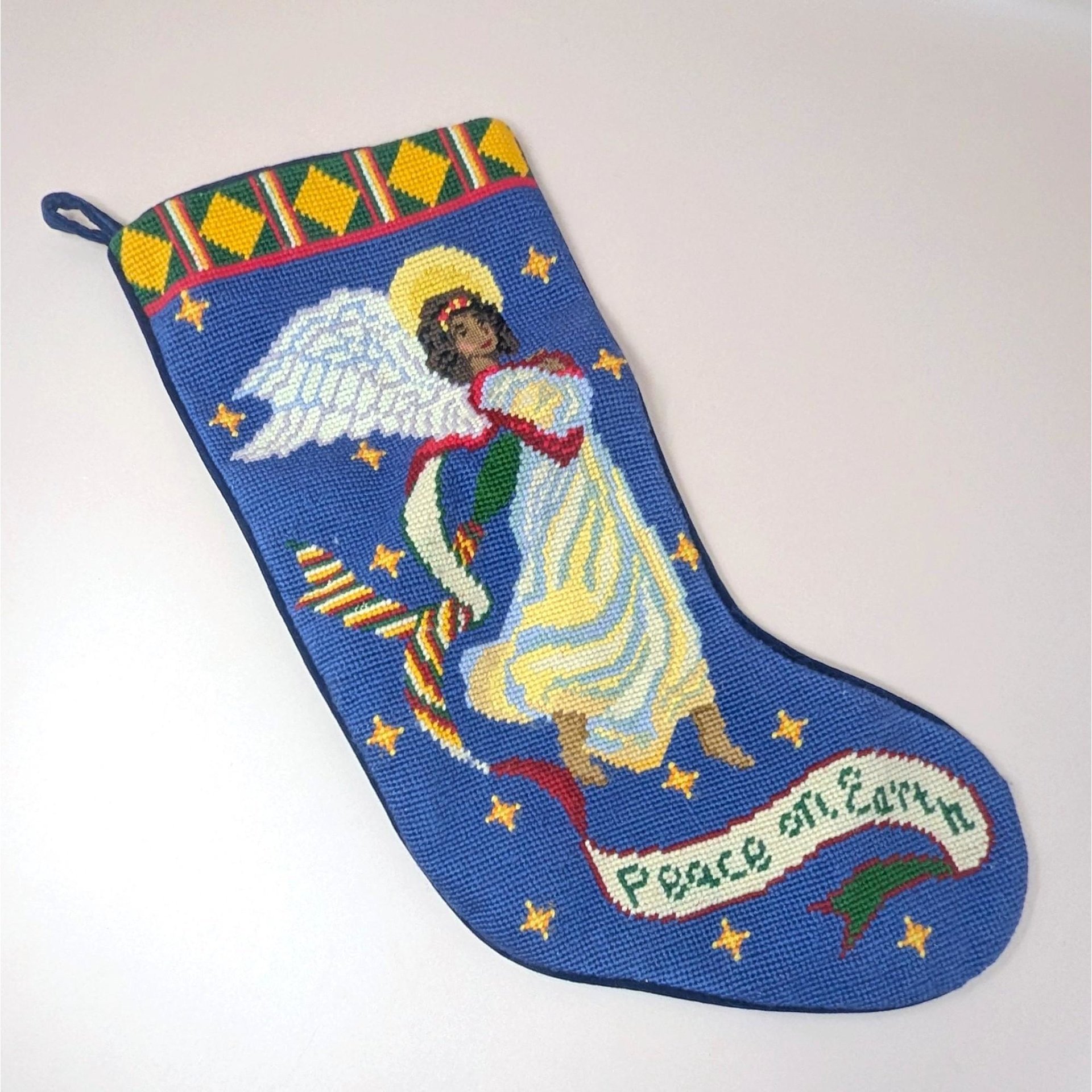 A vintage handmade needlepoint Christmas stocking on a soft pink background. The blue stocking features a hand stitched Black angel with white wings and a gold halo, surrounded by yellow stars and holding a banner reading Peace on Earth.