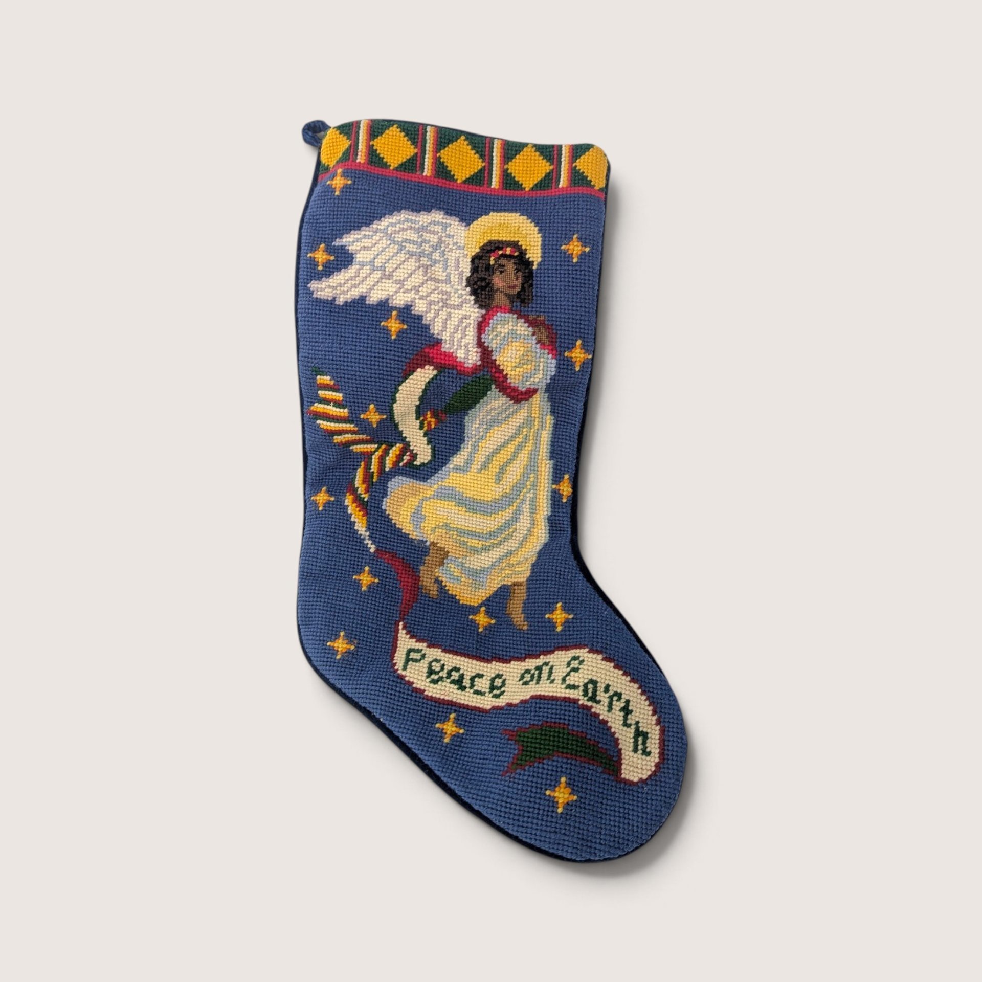 A vintage needlepoint Christmas stocking with a blue background featuring a winged angel with a halo, golden stars, and a banner that reads Peace on Earth.
