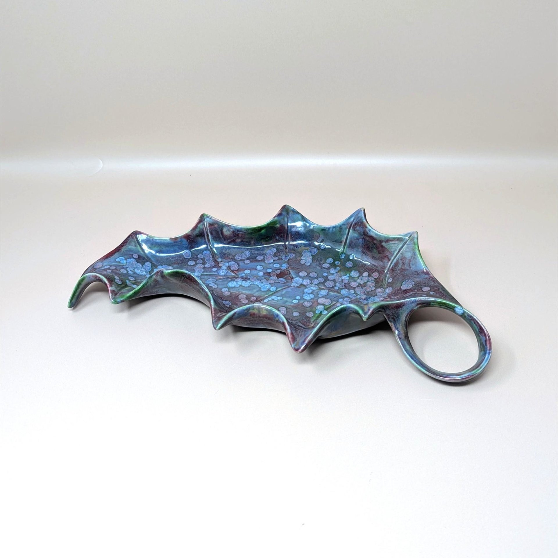 A large vintage studio art pottery leaf dish resting on a soft pink background. The ceramic serving tray is shaped like a stylized holly leaf with a loop handle, covered in a glossy, reactive drip glaze featuring speckled blue and deep purple tones.