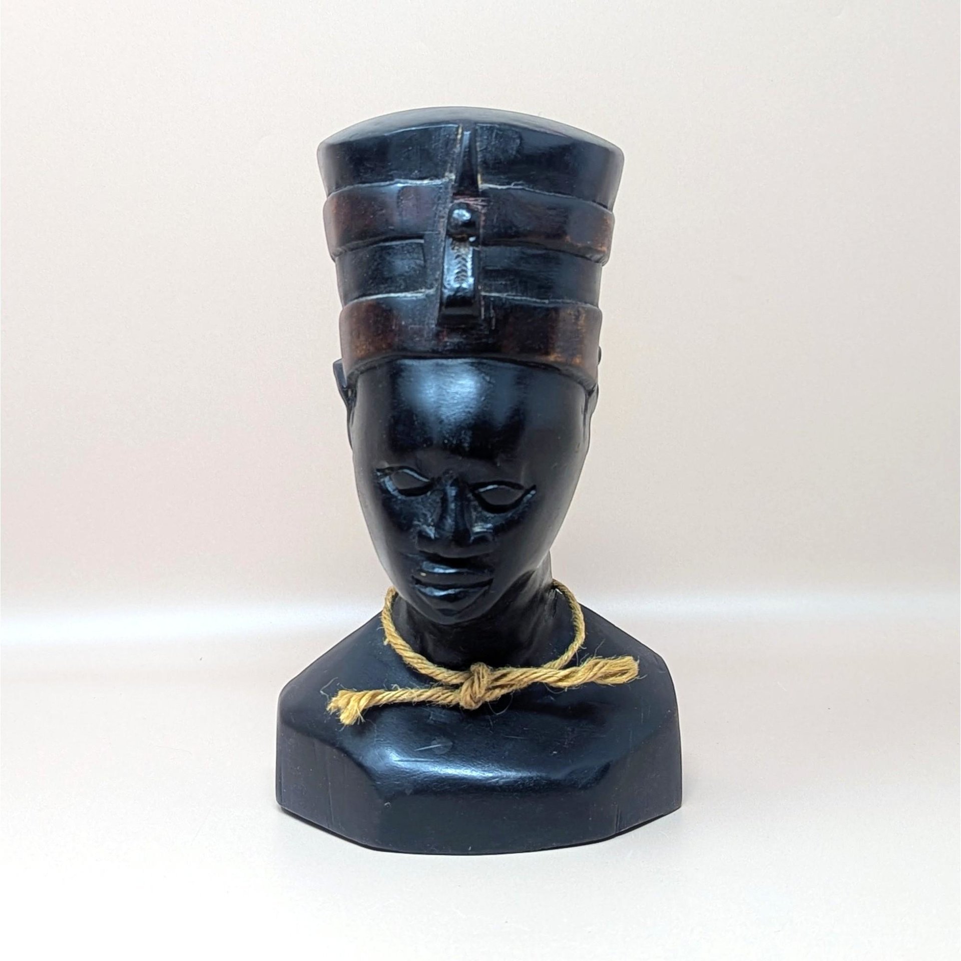 A vintage hand carved wooden bust of Egyptian Queen Nefertiti resting on a soft pink background. The dark polished sculpture features a tall flat topped crown with carved bands and a simple tan fiber rope knotted around the neck.